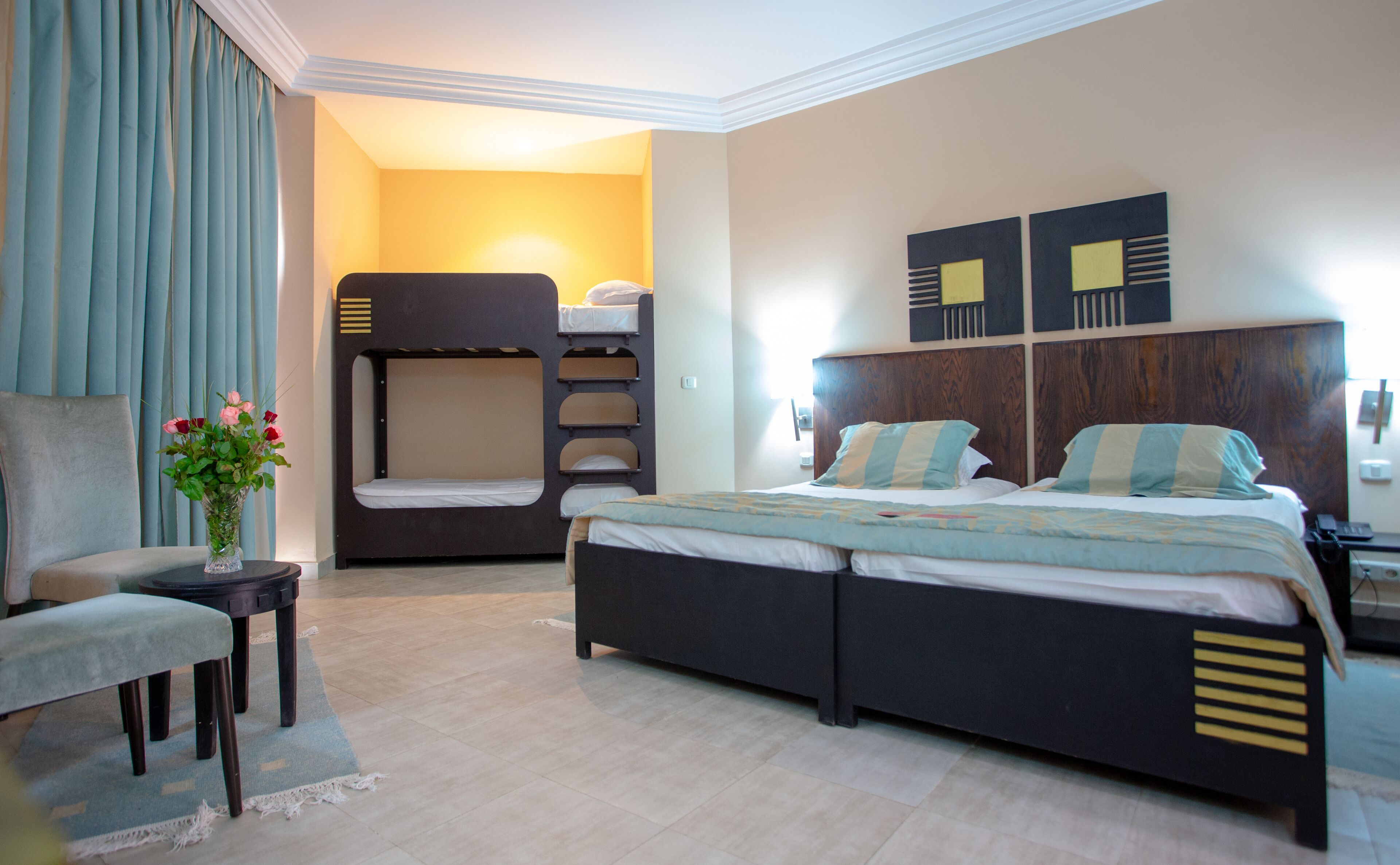 A hotel room with a bunk bed, a single bed, a chair, a small table with a vase of flowers, and a wall with two decorative panels.