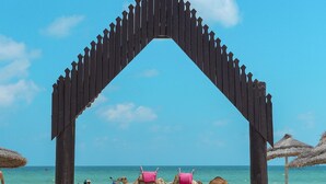 A wooden archway on a beach with a clear blue sky, leading to a sandy area with beach chairs and large clay pots. - Hotel Fiesta Beach Djerba (Djerba Midun)