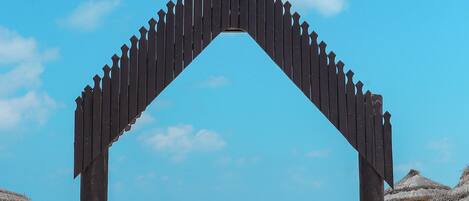 Un arco de madera en la playa bajo un cielo azul despejado, que conduce a una zona arenosa con sillas de playa y grandes macetas de barro.