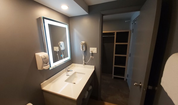 A modern bathroom with a white sink, two hair dryers mounted on the wall, and a mirror with a lighted frame.