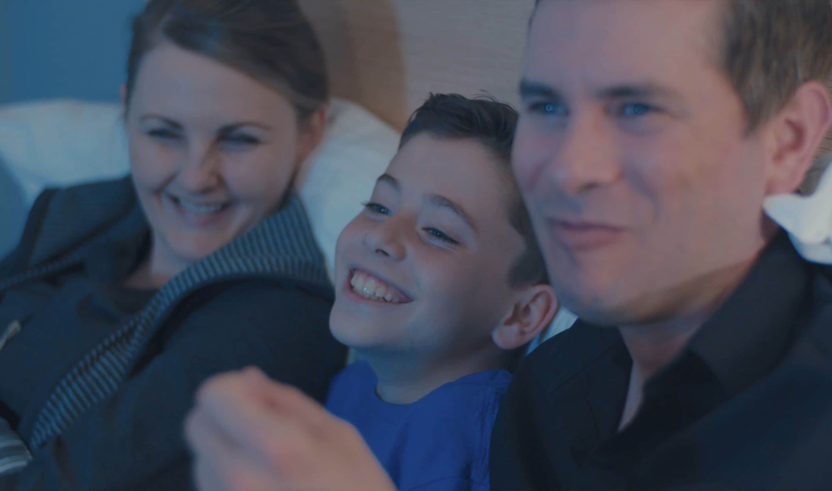 Una familia de tres en una habitación con poca luz, probablemente de un hotel, con una cama y una ventana al fondo.