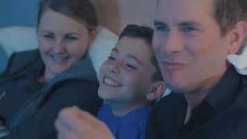 Una familia de tres en una habitación con poca luz, probablemente de un hotel, con una cama y una ventana al fondo.