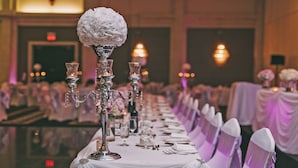 Un salón de banquetes con una larga mesa dispuesta para un evento formal, decorada con candelabros de cristal y arreglos florales blancos.