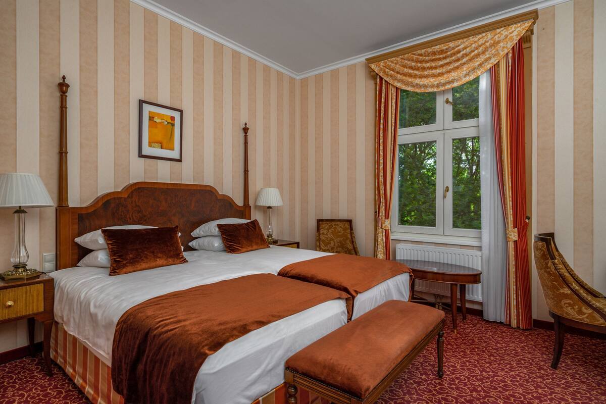 Ein Doppelbett mit Holz-Kopfteil, zwei Nachttische mit Lampen, ein Stuhl und ein Fenster mit Vorhängen.
