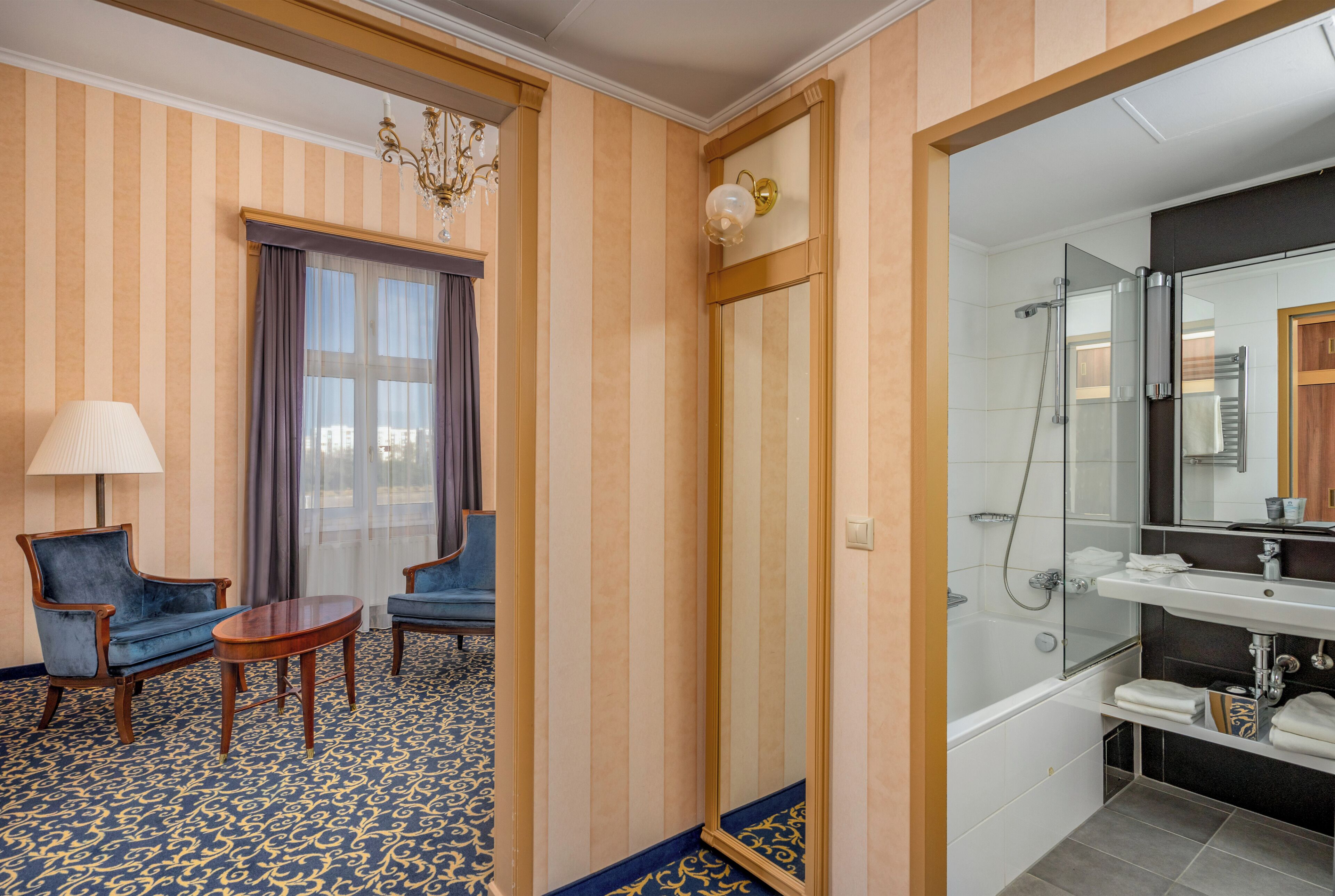 Una habitación de hotel con papel tapiz a rayas, una araña, dos sillones, una mesita, un espejo grande, una bañera y un lavabo.