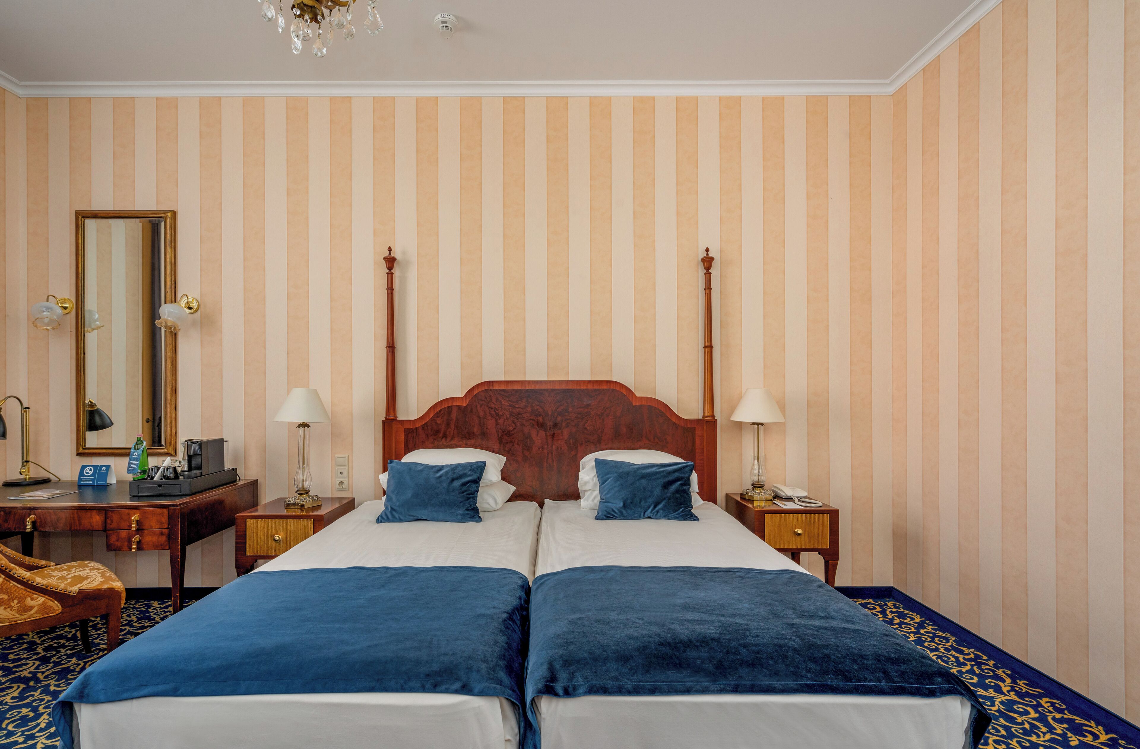 A hotel room with two beds, striped wallpaper, a wooden headboard, bedside tables with lamps, and a large mirror.