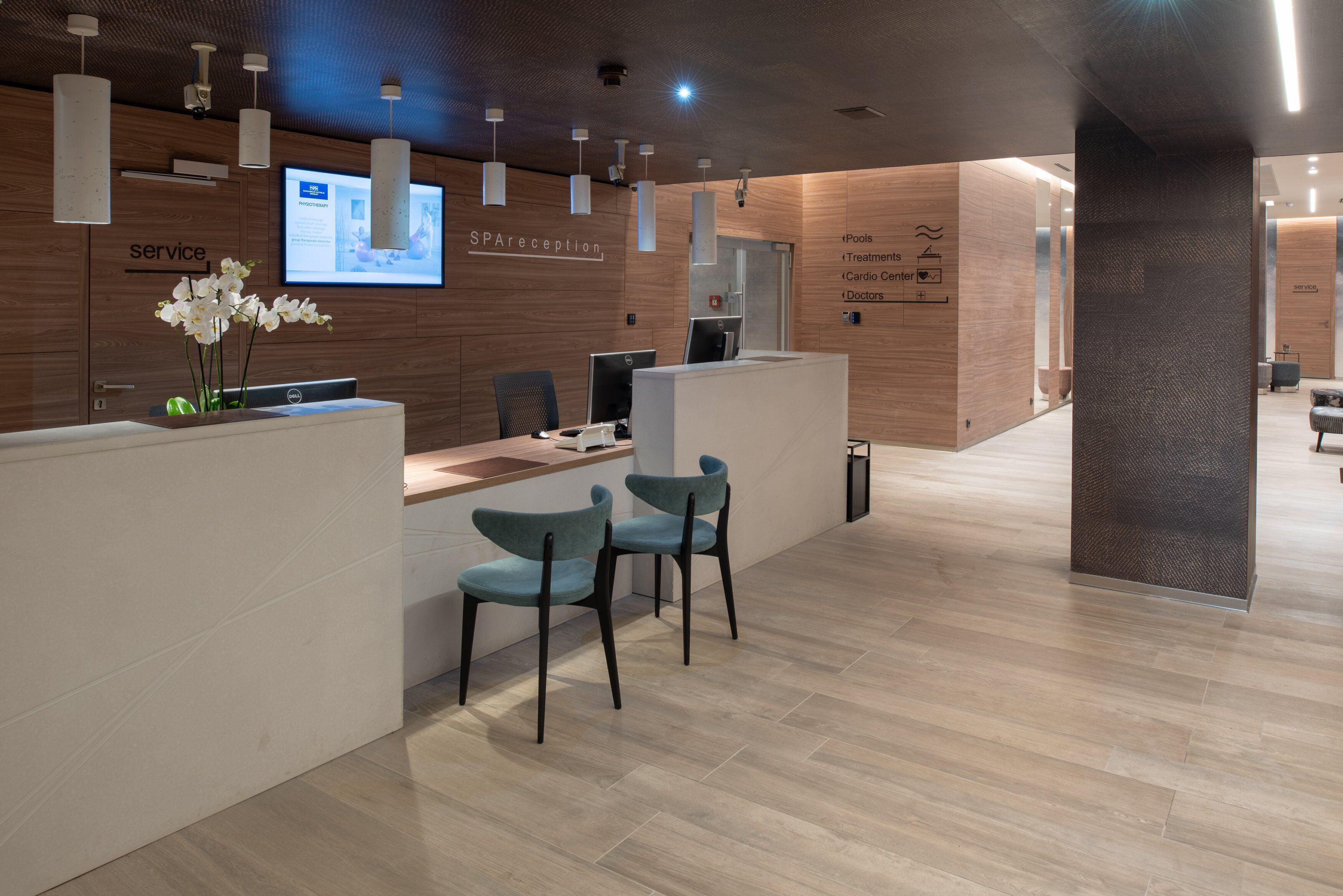 A reception area with a wooden reception desk, two blue chairs, a computer, and a screen displaying information.