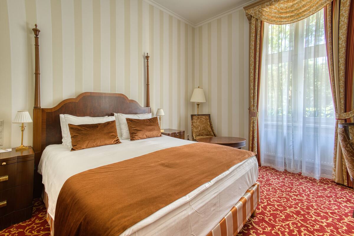 Ein Hotelzimmer mit einem großen Bett, einem Nachttisch, einer Lampe, einem Sessel und einem Fenster mit Vorhängen.