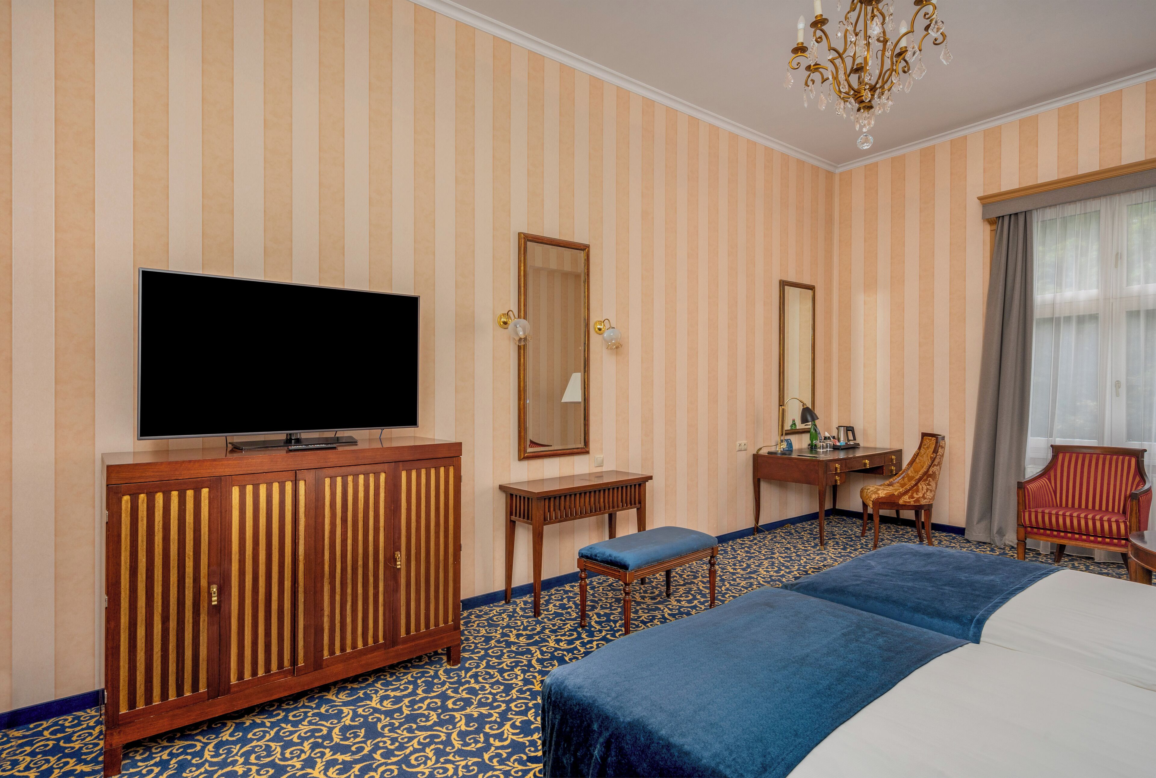 Quarto de hotel com uma cama grande, uma televisão sobre um móvel de madeira, uma escrivaninha com uma cadeira e um espelho amplo.