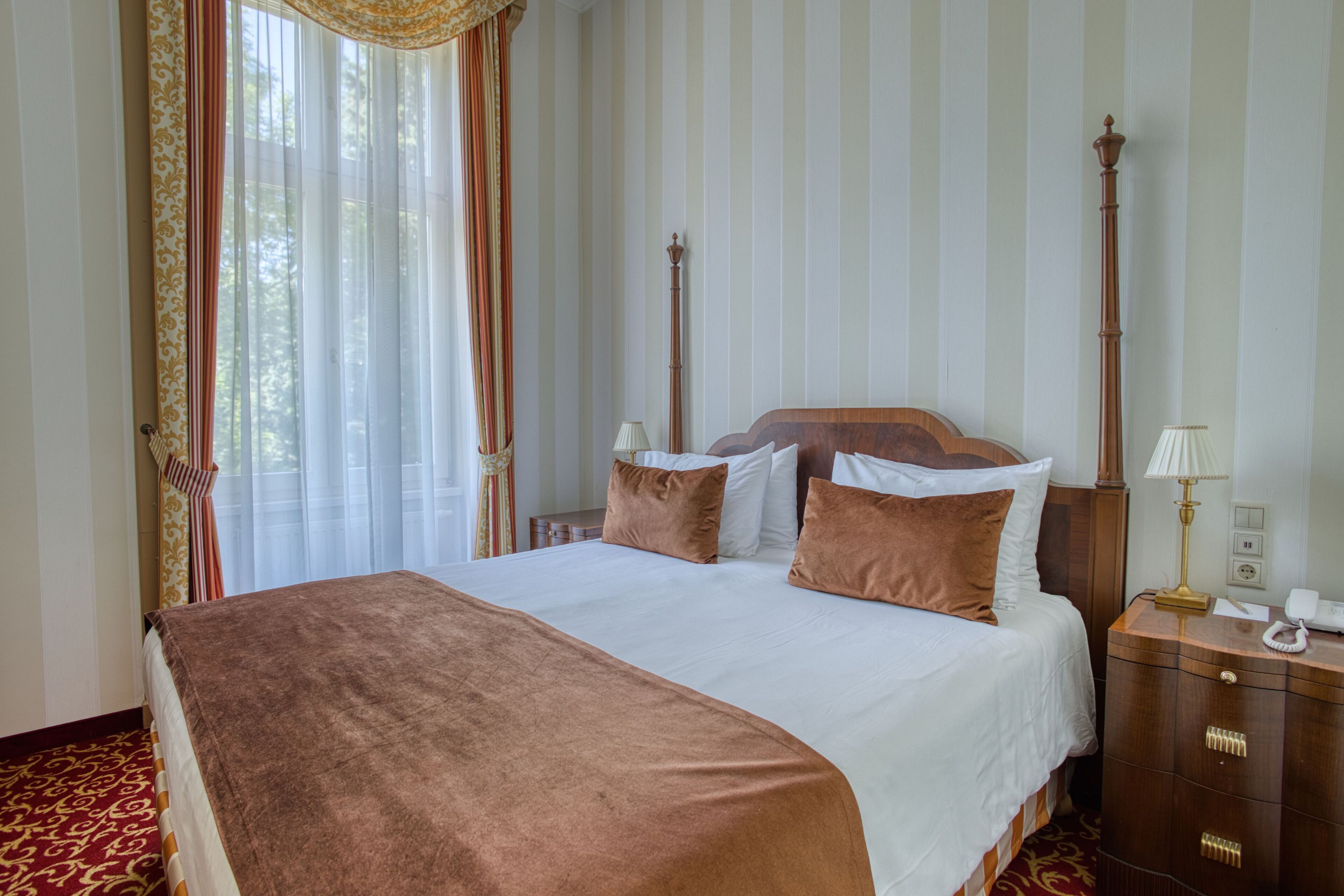 A large bed with a brown blanket, white pillows, and a wooden headboard. There is a nightstand with a lamp and a telephone. The room has striped wallpaper and a window with curtains.