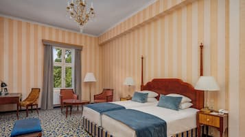 A hotel room with striped wallpaper, a large bed with two pillows, a nightstand, a chair, and a desk.