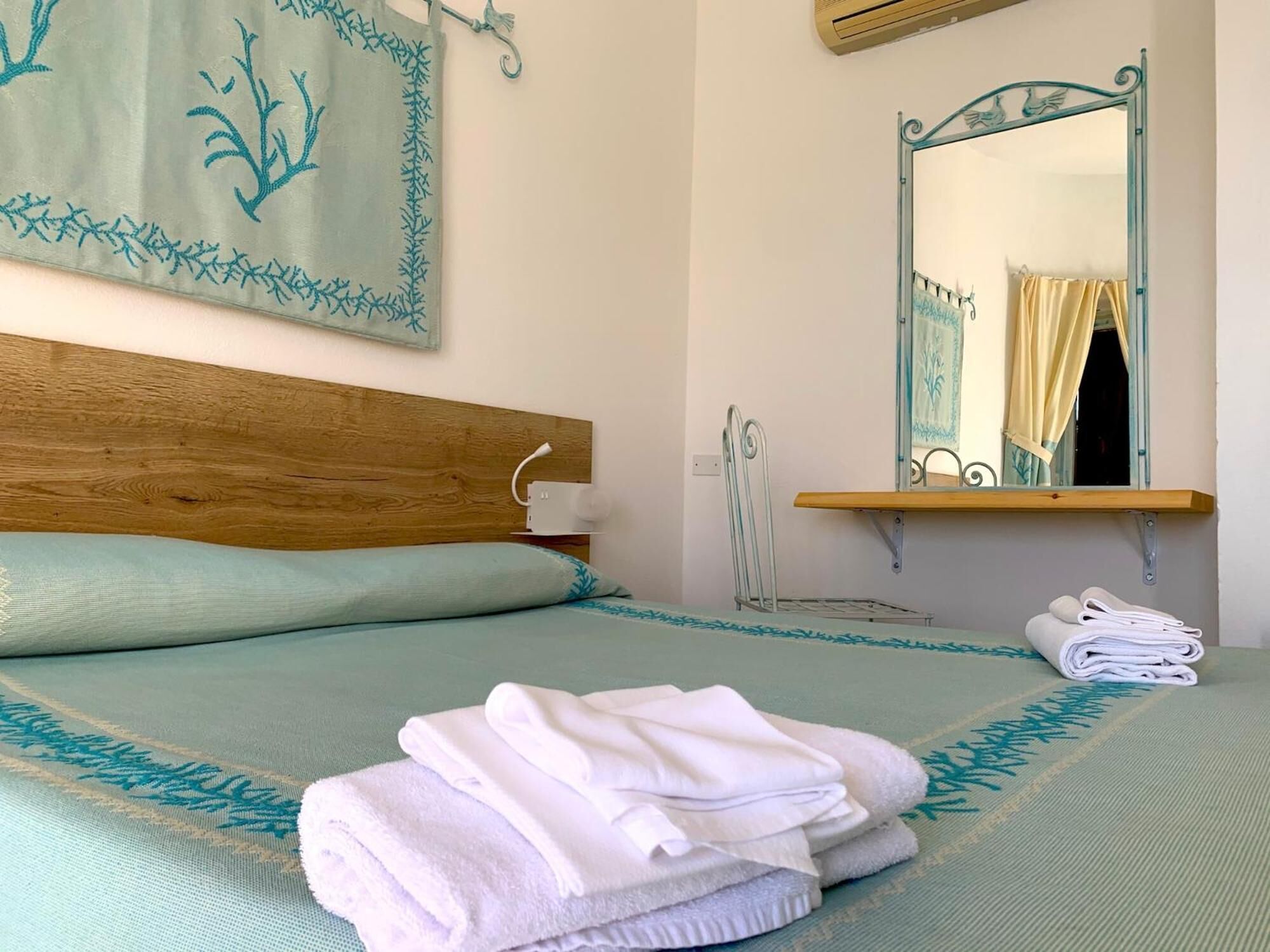 Een opgemaakt bed met een houten hoofdbord, een nachtkastje met een lamp, een spiegel met decoratieve lijst en een opgevouwen handdoek op het bed.