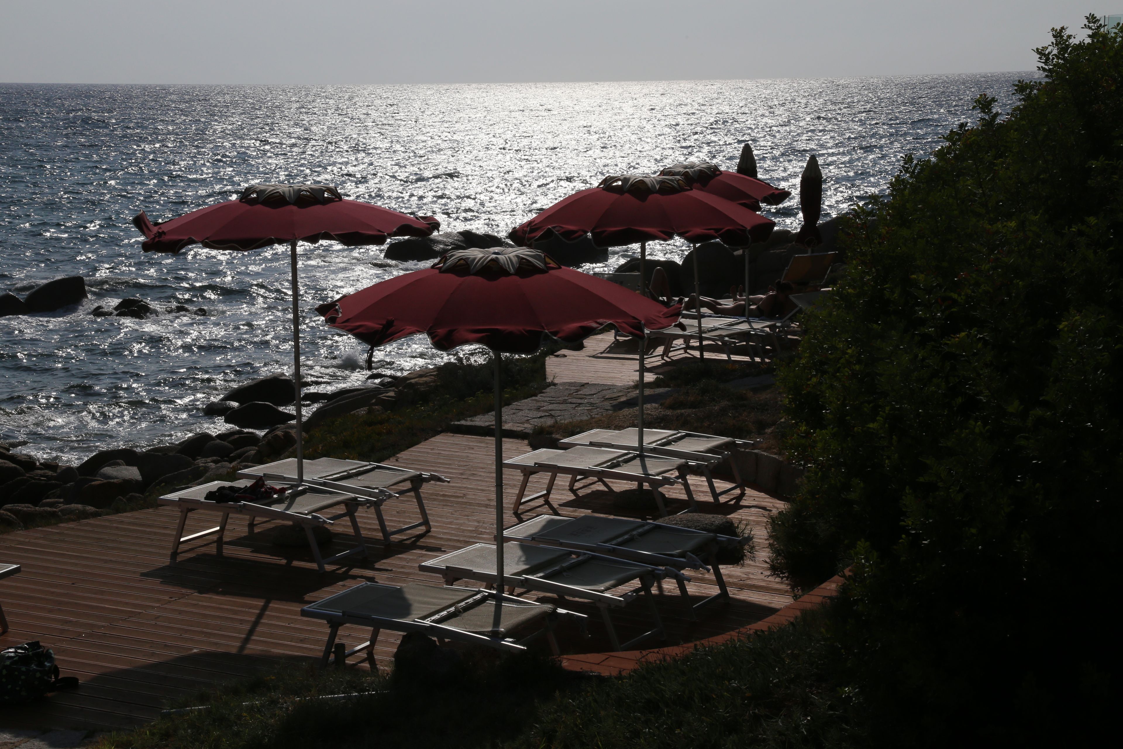 A seaside terrace with sun loungers and red umbrellas, overlooking the ocean.