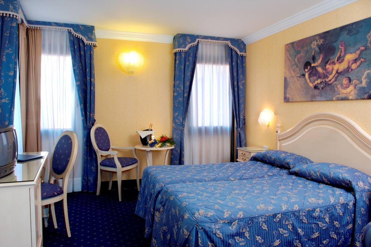 A hotel room with a bed, a desk with a computer, a chair, and a small table with a vase of flowers.
