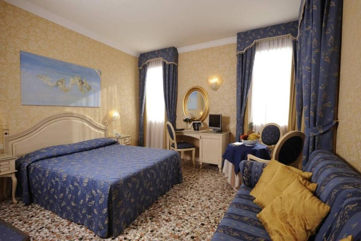 A hotel room with a large bed, a desk with a computer, a chair, a small table with a plant, and a sofa with cushions.