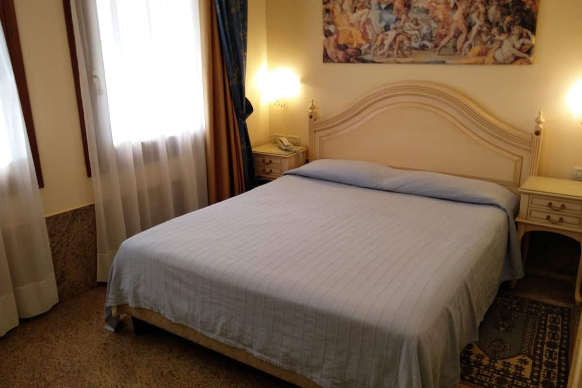 A hotel room with a bed, bedside tables, a painting on the wall, and a window with curtains.