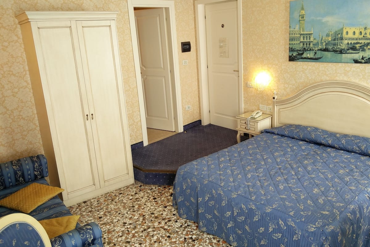 A hotel room with a bed, a wardrobe, a desk with a lamp, a painting of a cityscape, and a patterned carpet.