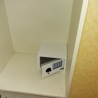 A white minibar with a digital lock and a small display screen, located in a corner of a room with plain walls.