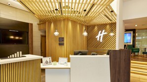 A reception area with a wooden reception desk, a mounted TV, and a sign with a large 'H'. - Holiday Inn Newcastle Jesmond by IHG (Newcastle-upon-Tyne)