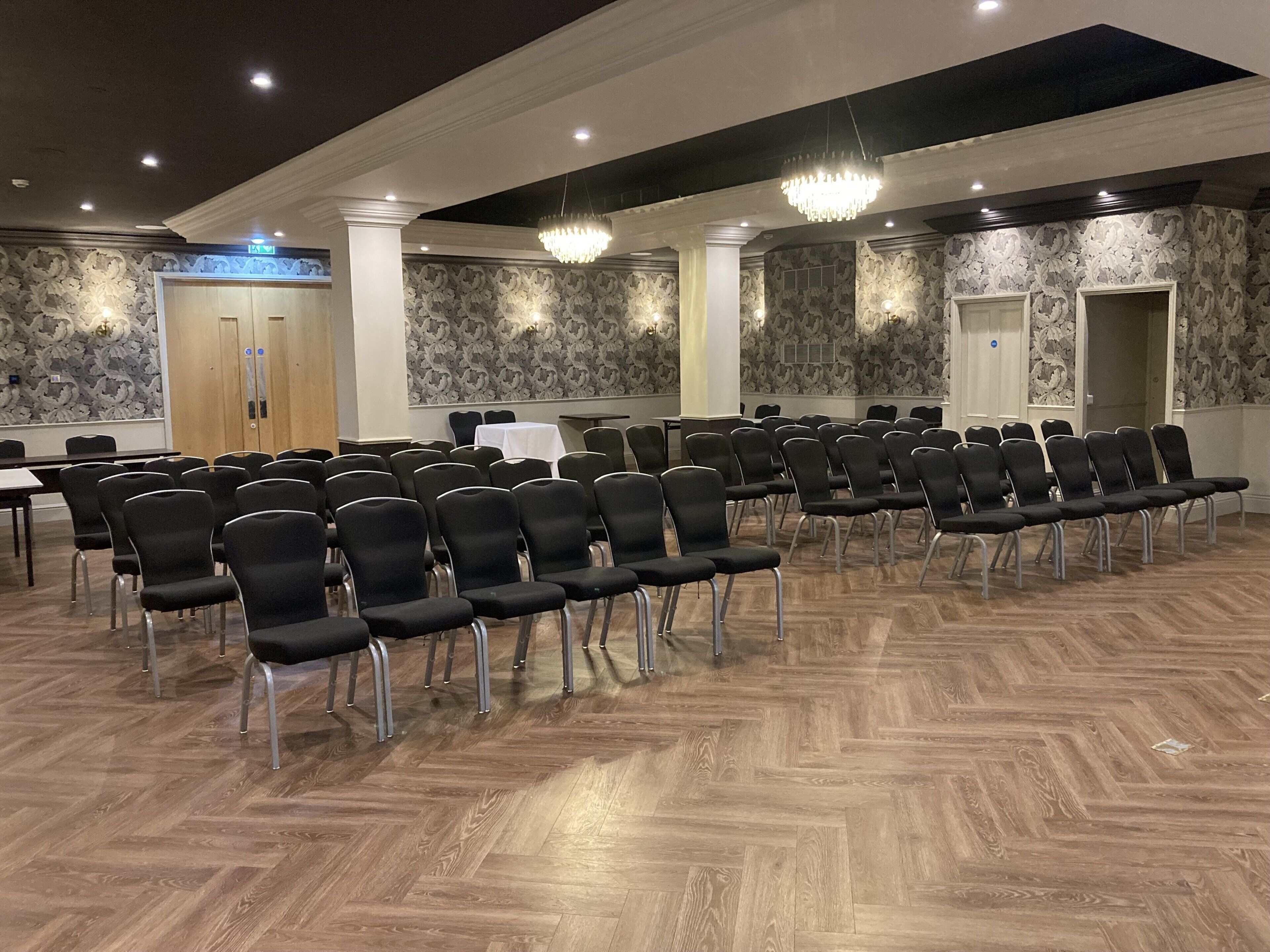 Konferenssihuone, jossa on riveissä mustia tuoleja, valkoinen pöytä ja kuviollinen tapetti.