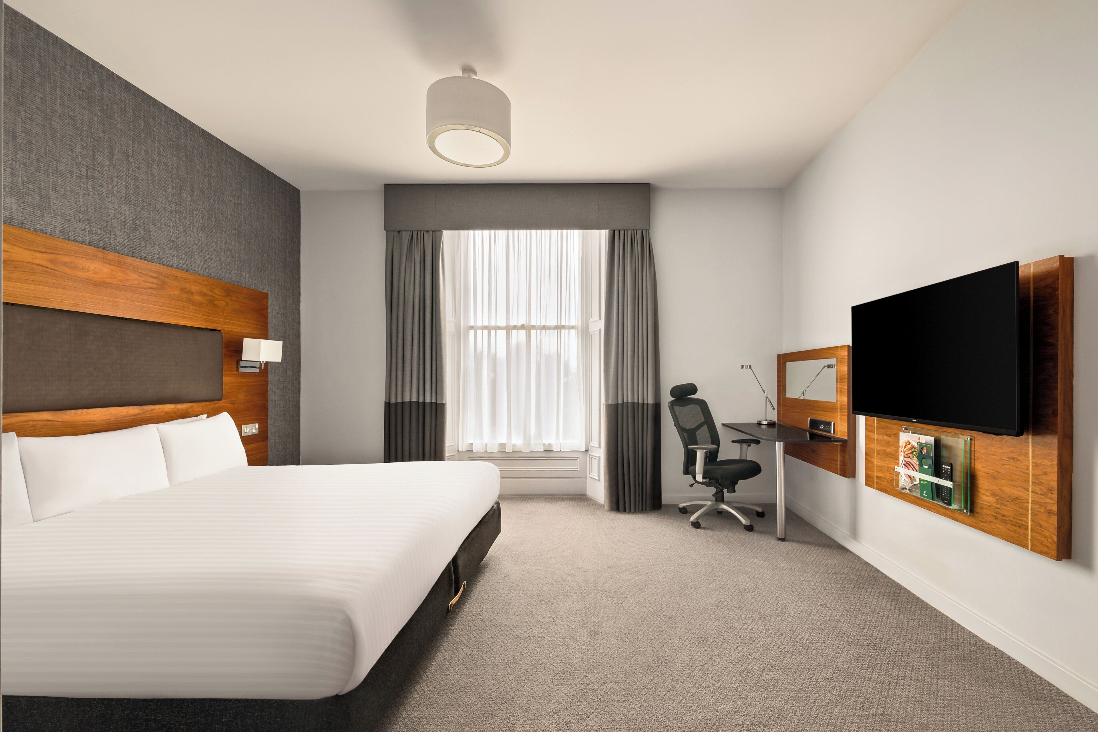 Une chambre d’hôtel avec un grand lit, un bureau avec une chaise, un téléviseur à écran plat fixé au mur et une fenêtre avec des rideaux.