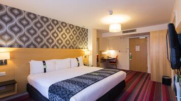 A hotel room with a large bed, a desk, a chair, a television, and a patterned wall.
