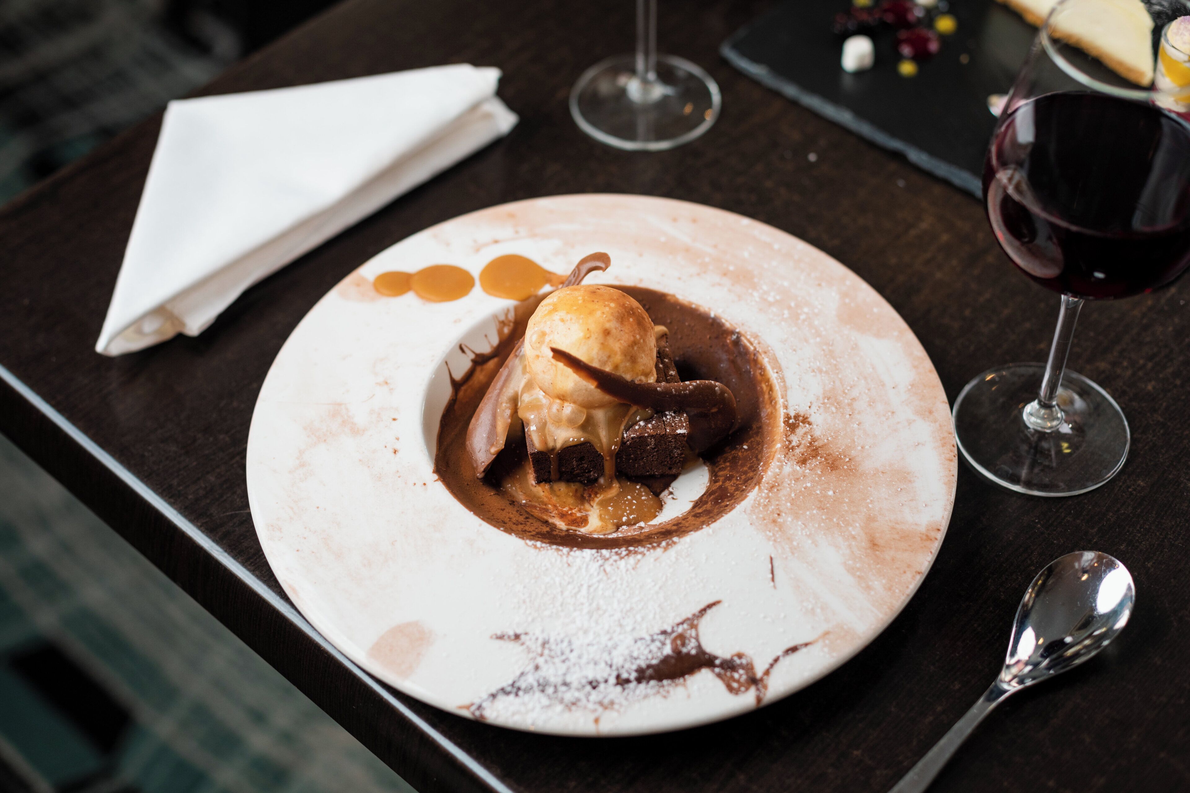 A dessert plate with a scoop of ice cream and a chocolate piece, accompanied by a wine glass and a spoon on a table.