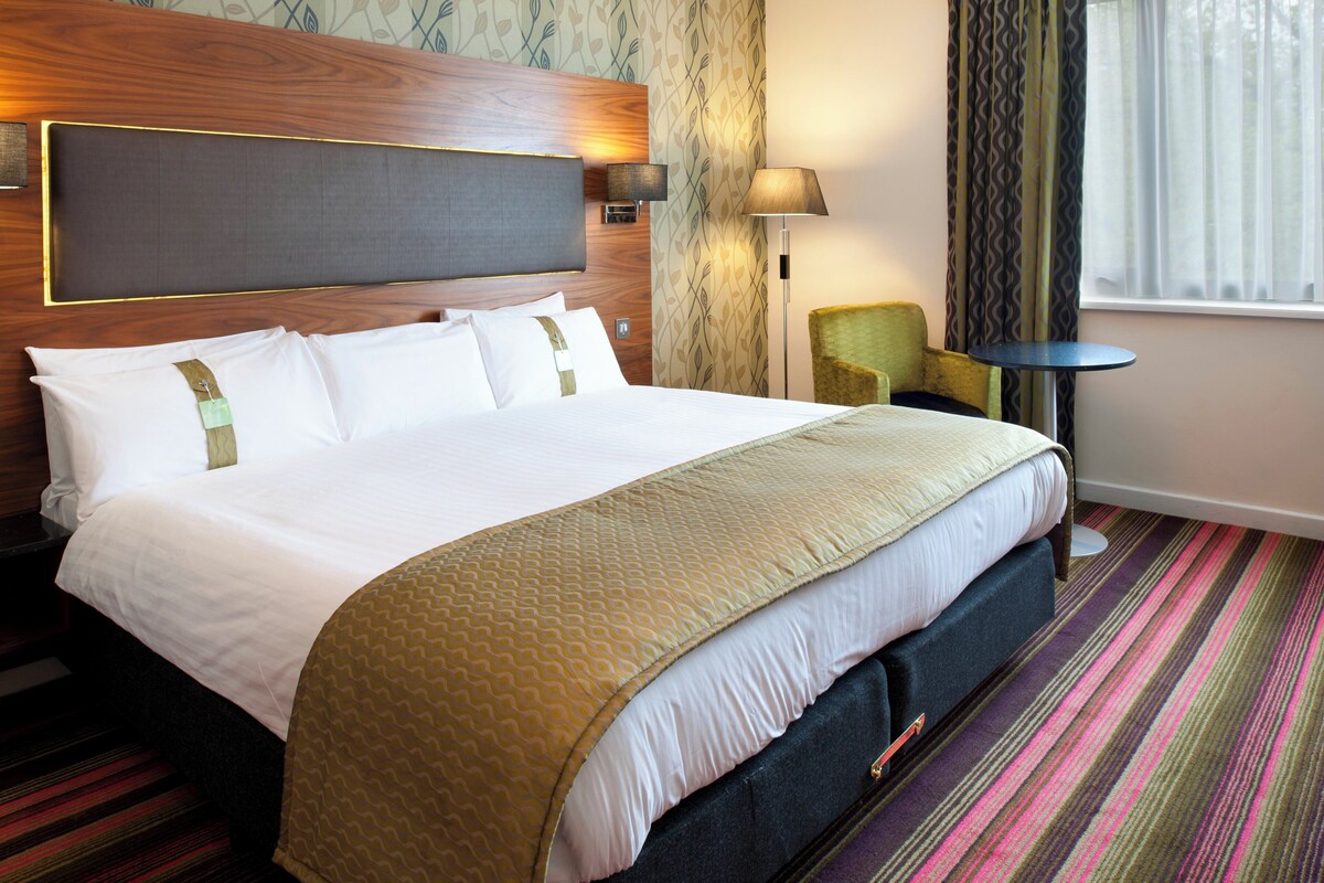 A hotel room with a large bed, a headboard, a bedside table, a lamp, a chair, and a window with curtains.