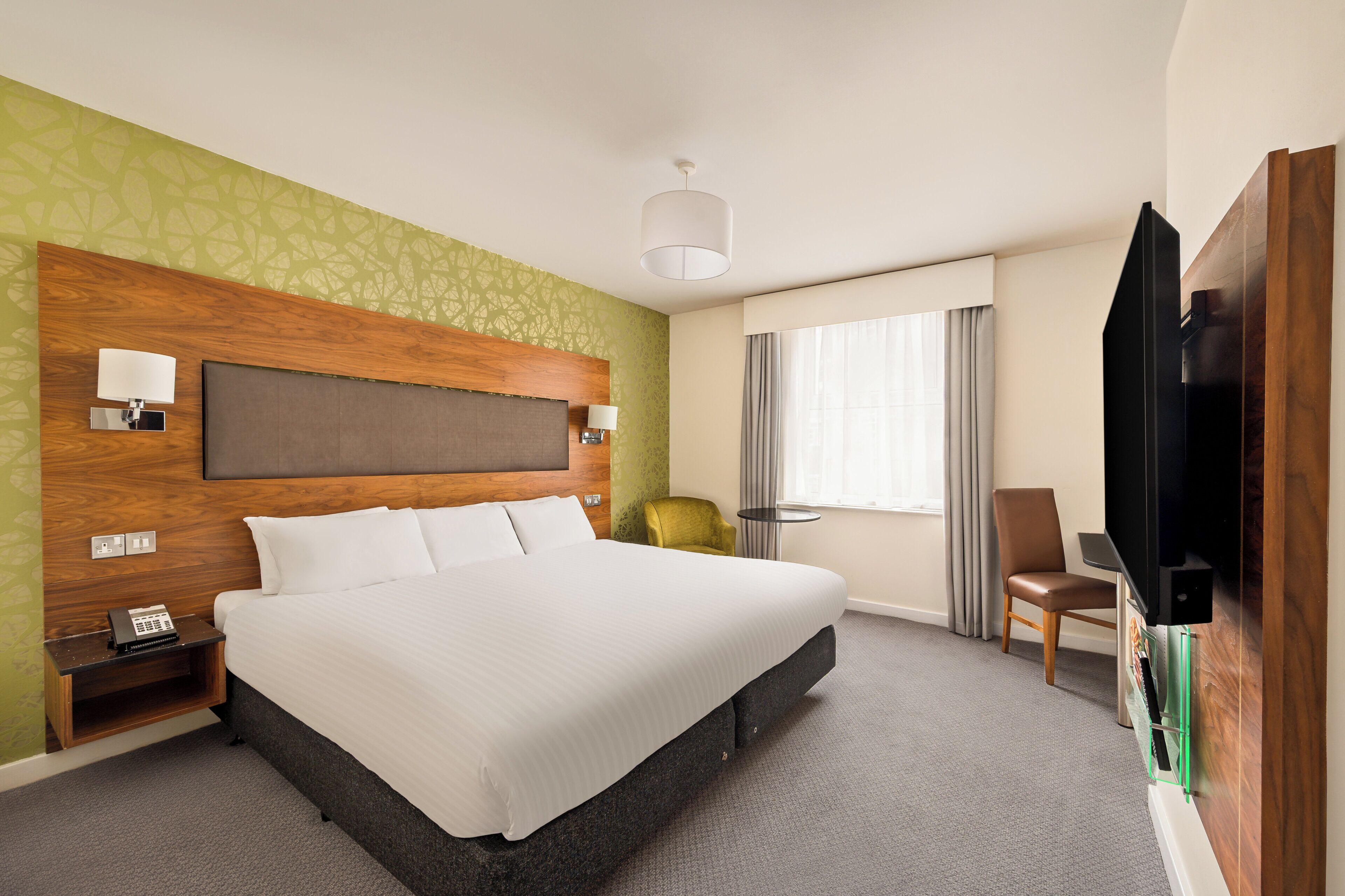 Een hotelkamer met een groot bed, een houten hoofdbord, een nachtkastje met een lamp, een stoel, een televisie en een raam met gordijnen.