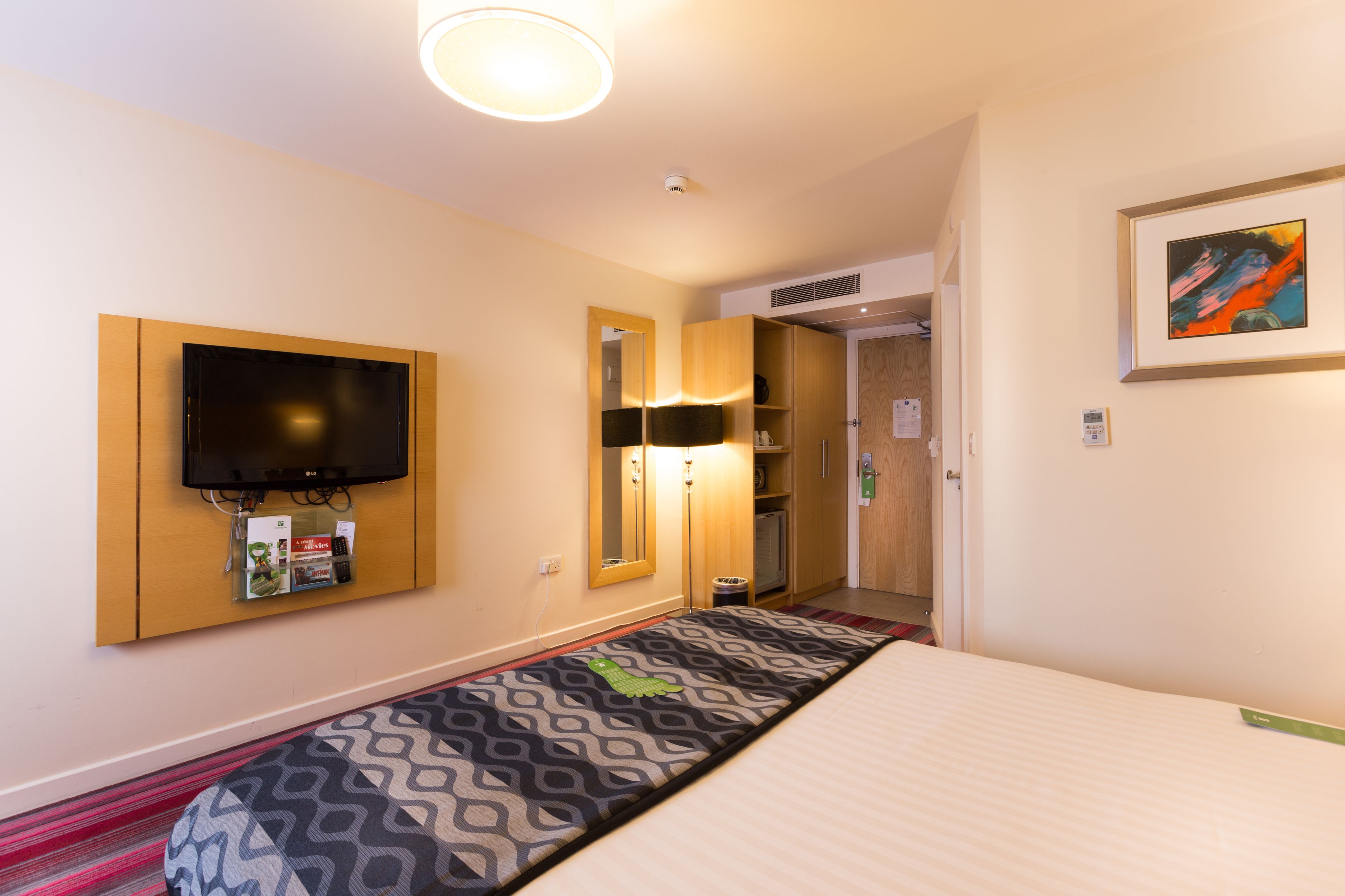 A hotel room with a bed, a television mounted on the wall, a mirror, a shelf with toiletries, and a framed picture on the wall.