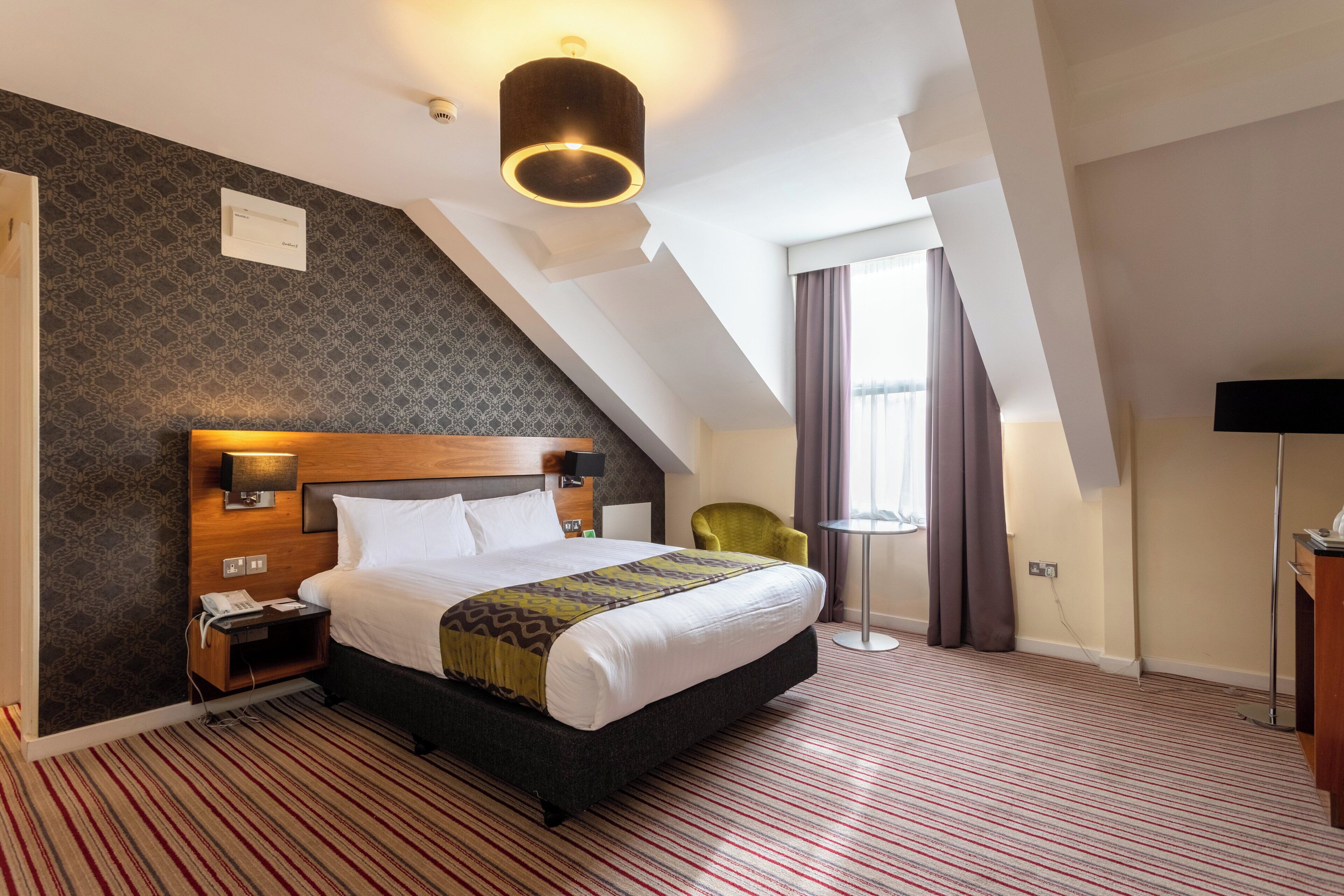 A hotel room with a large bed, a wooden headboard, a bedside table with a lamp, a small desk, a chair, and a large window with curtains.