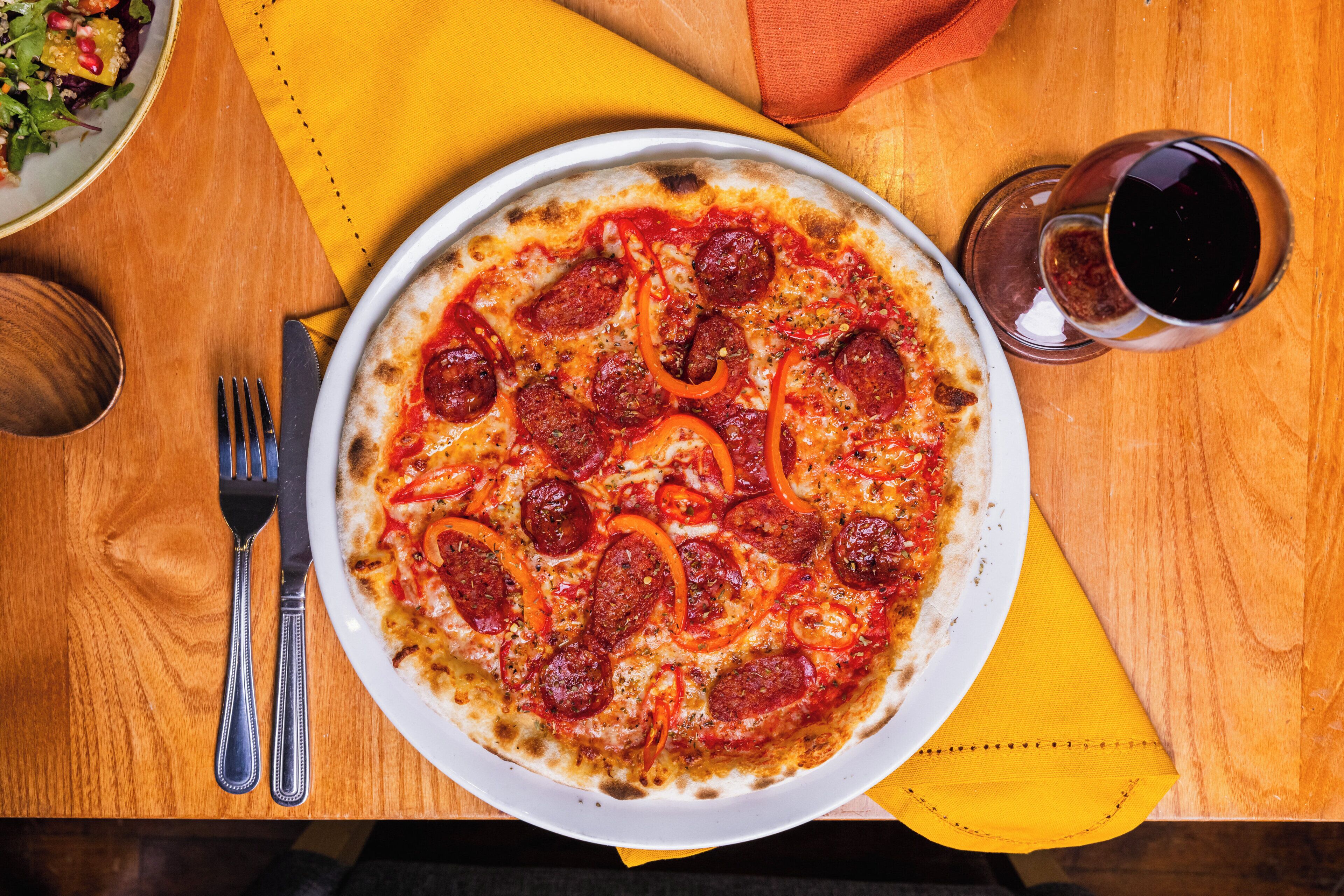 A pizza with pepperoni and red bell peppers on a white plate, placed on a wooden table with a yellow napkin and a glass of red wine.