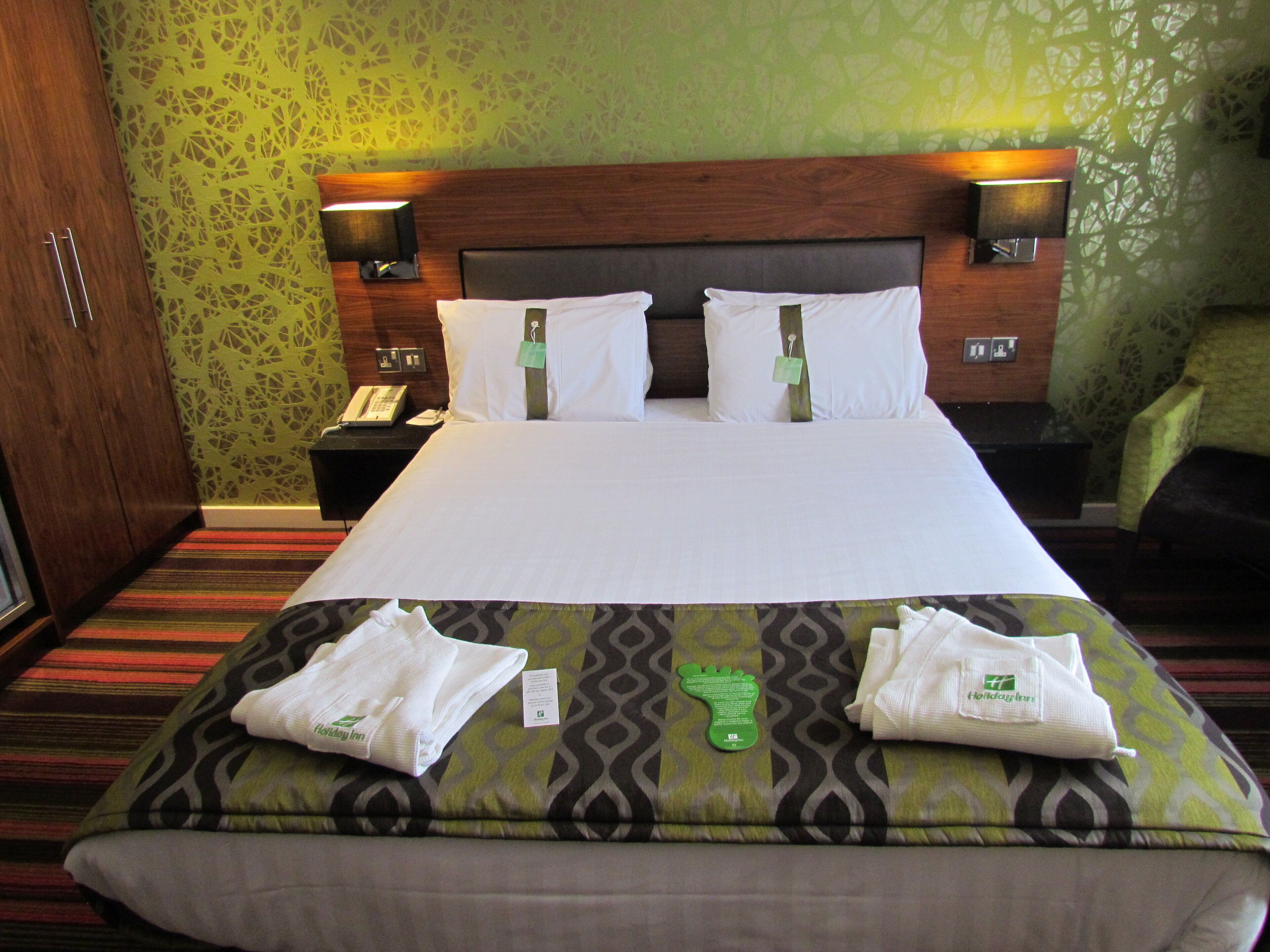A hotel room with a large bed, two bedside tables, a nightstand, a green patterned wall, and a green patterned sofa.