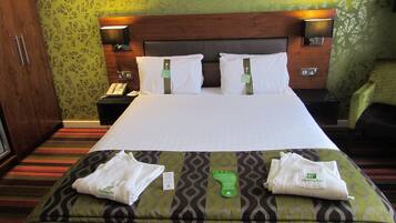 A hotel room with a large bed, two bedside tables, a nightstand, a green patterned wall, and a green patterned sofa.