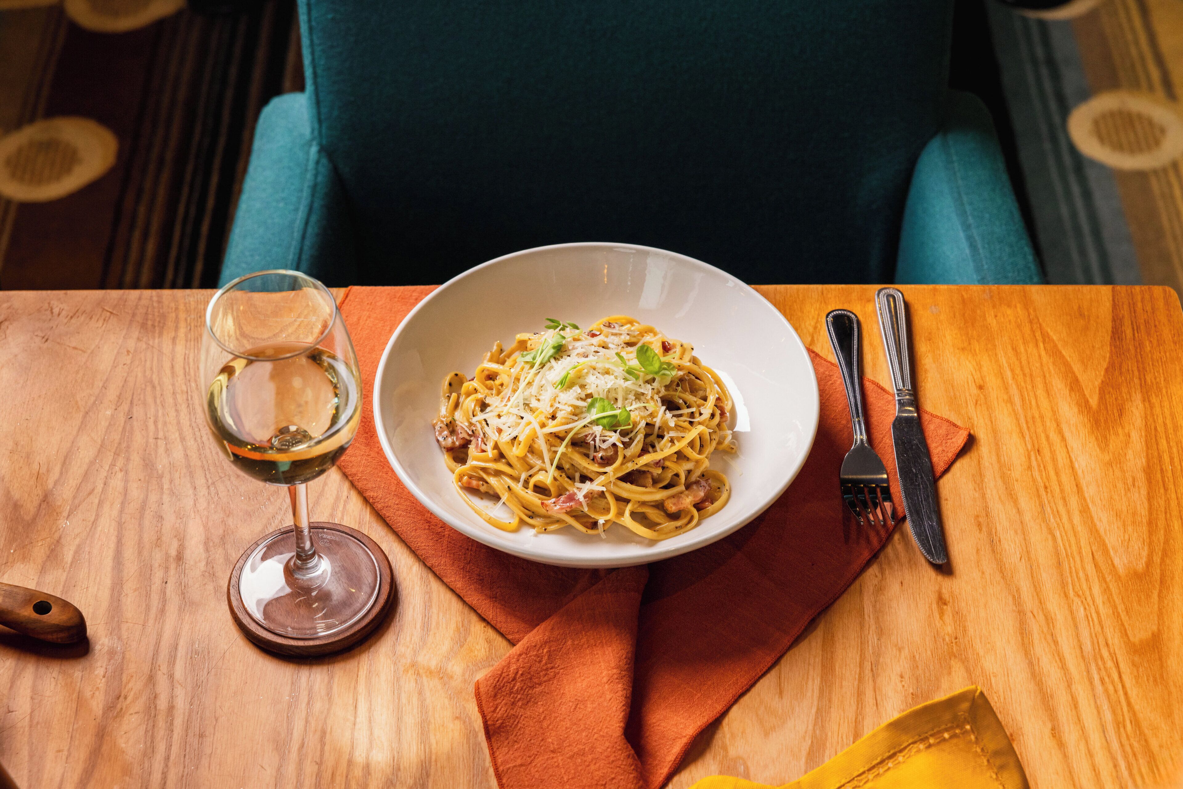 A wooden table with a bowl of pasta, a wine glass, and cutlery.