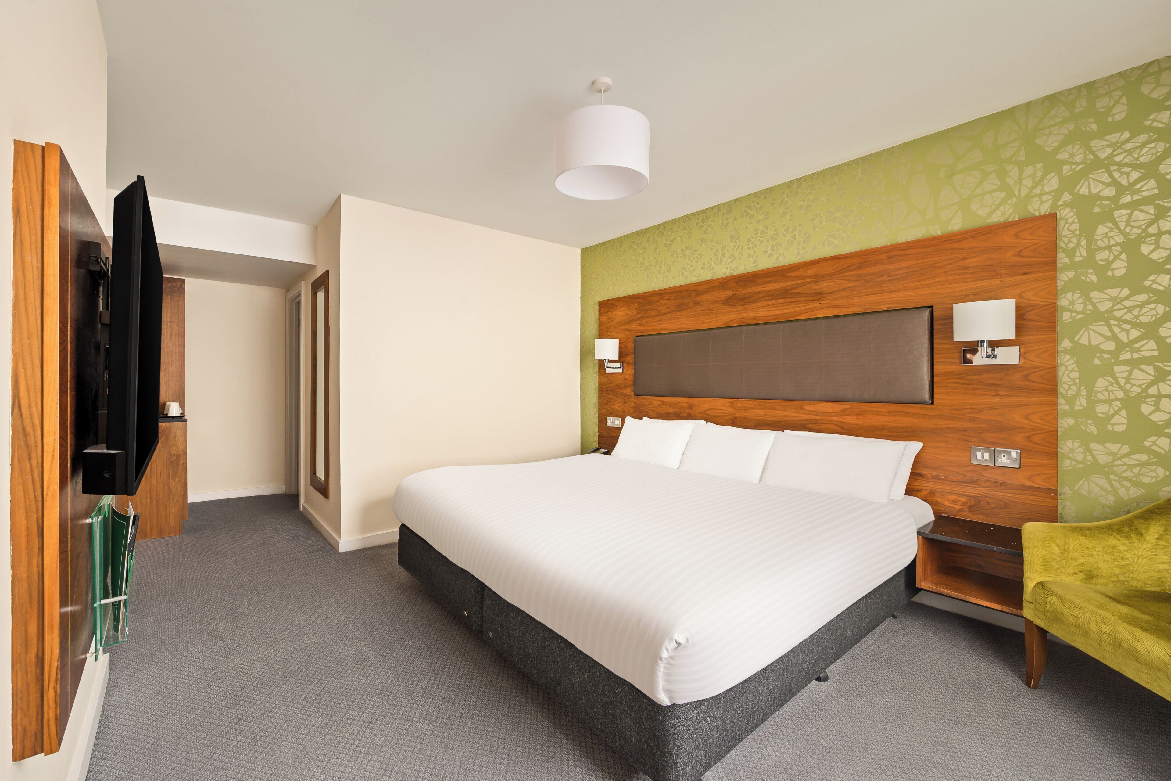 Habitación de hotel con una cama grande, cabecera de madera, televisión y una pared verde decorativa.