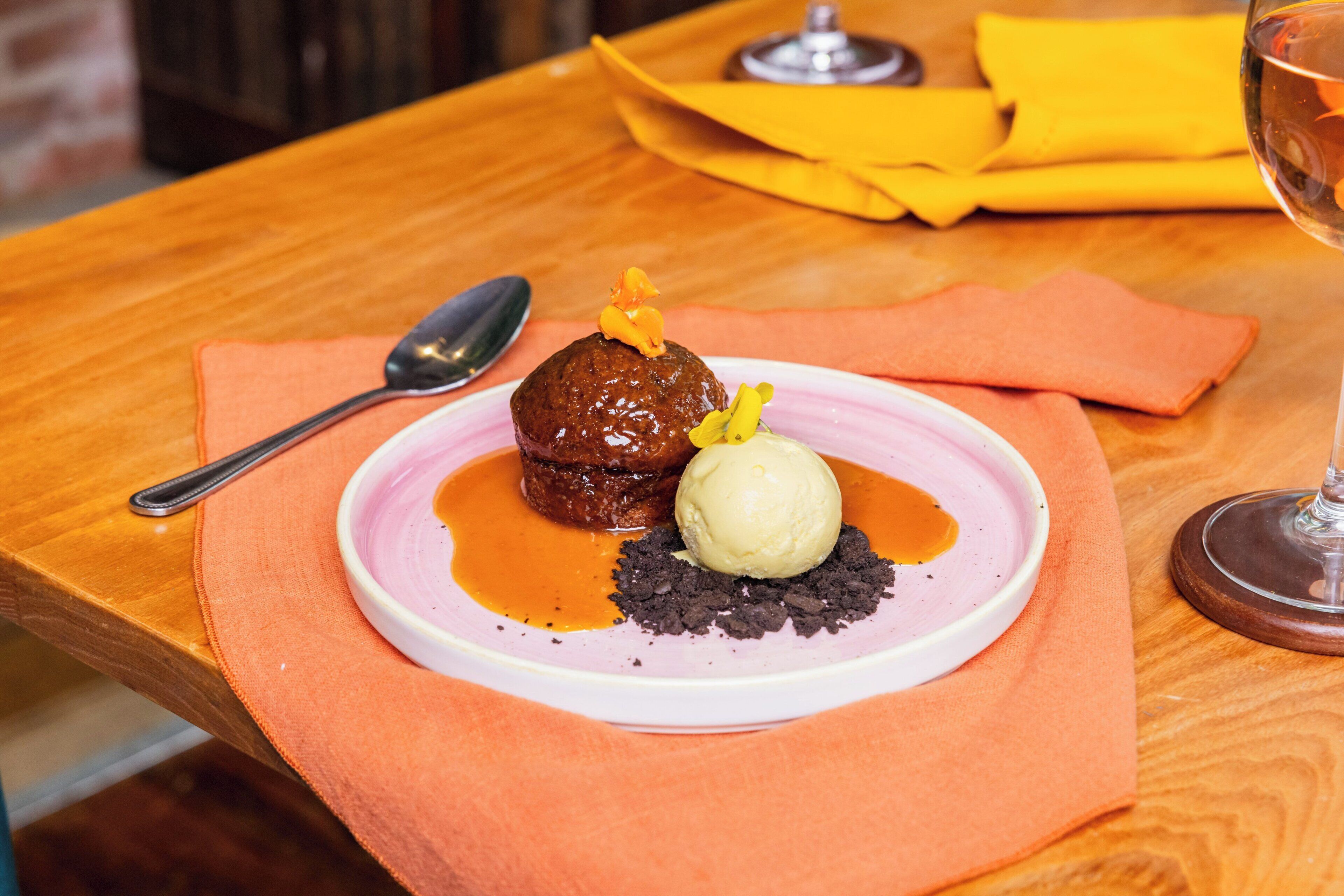 A dessert plate with a chocolate lava cake, a scoop of vanilla ice cream, and chocolate crumble on a pink plate, placed on an orange napkin.