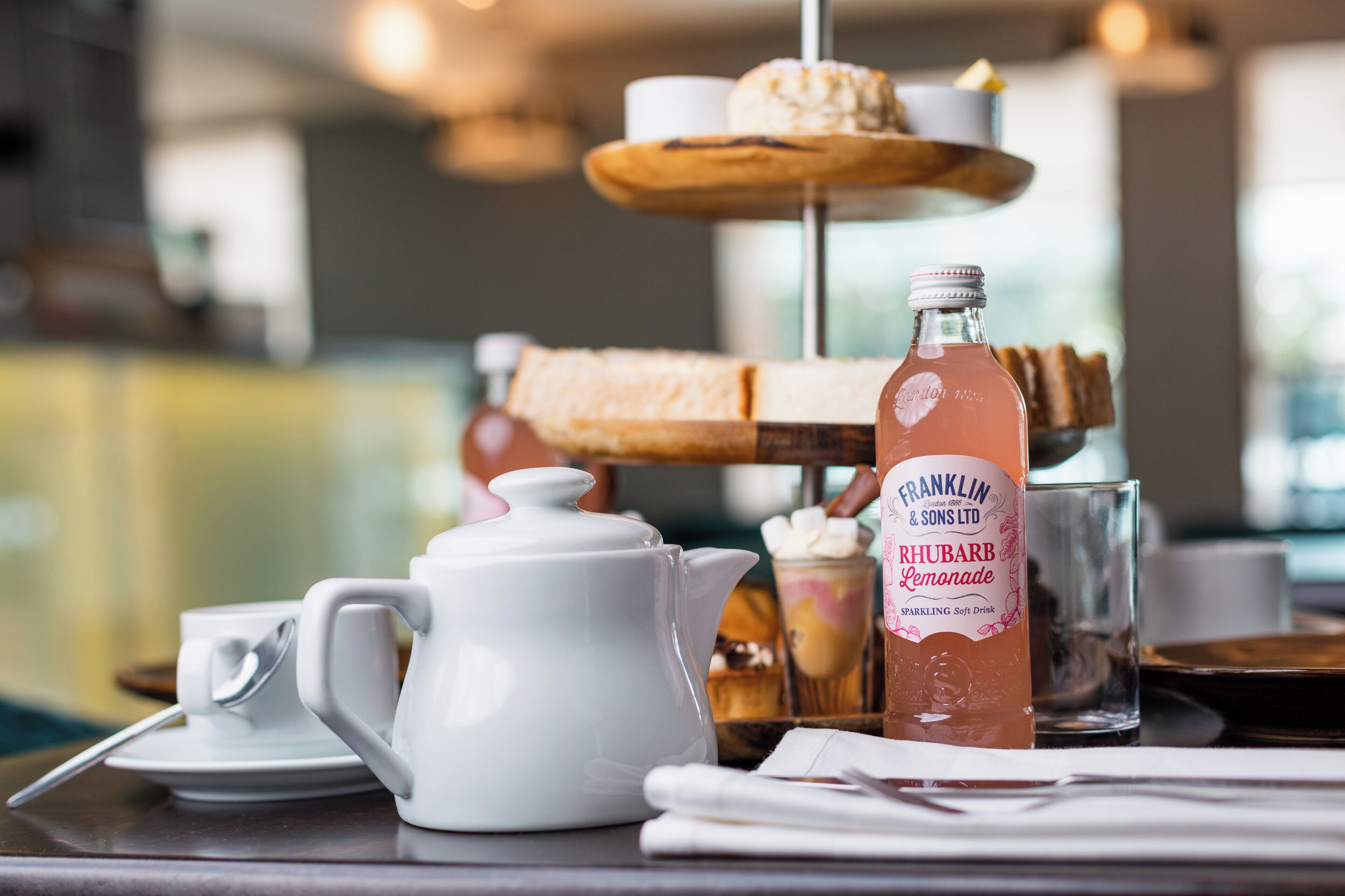 A table with a teapot, a bottle of lemonade, a glass, and a dessert display.