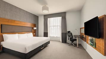 A hotel room with a large bed, a desk with a chair, a flat-screen TV, and a large window with curtains.