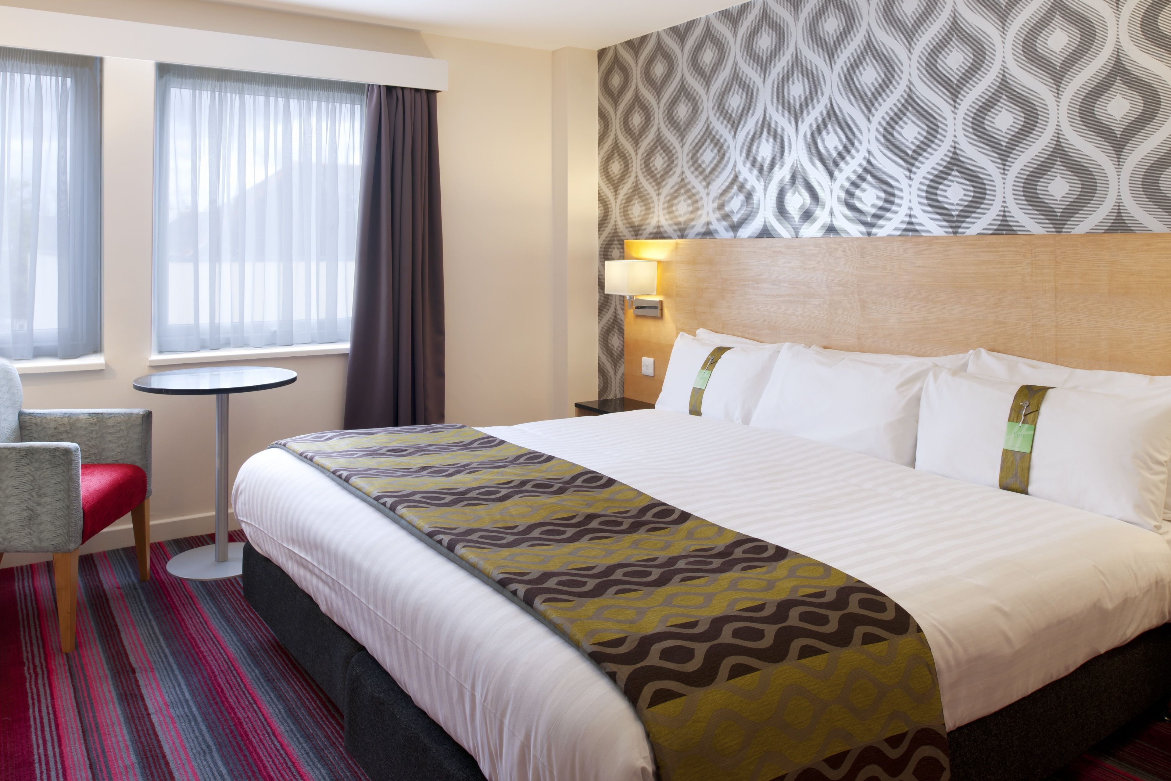 A hotel room with a large bed, a small round table, a chair, and a patterned wall.