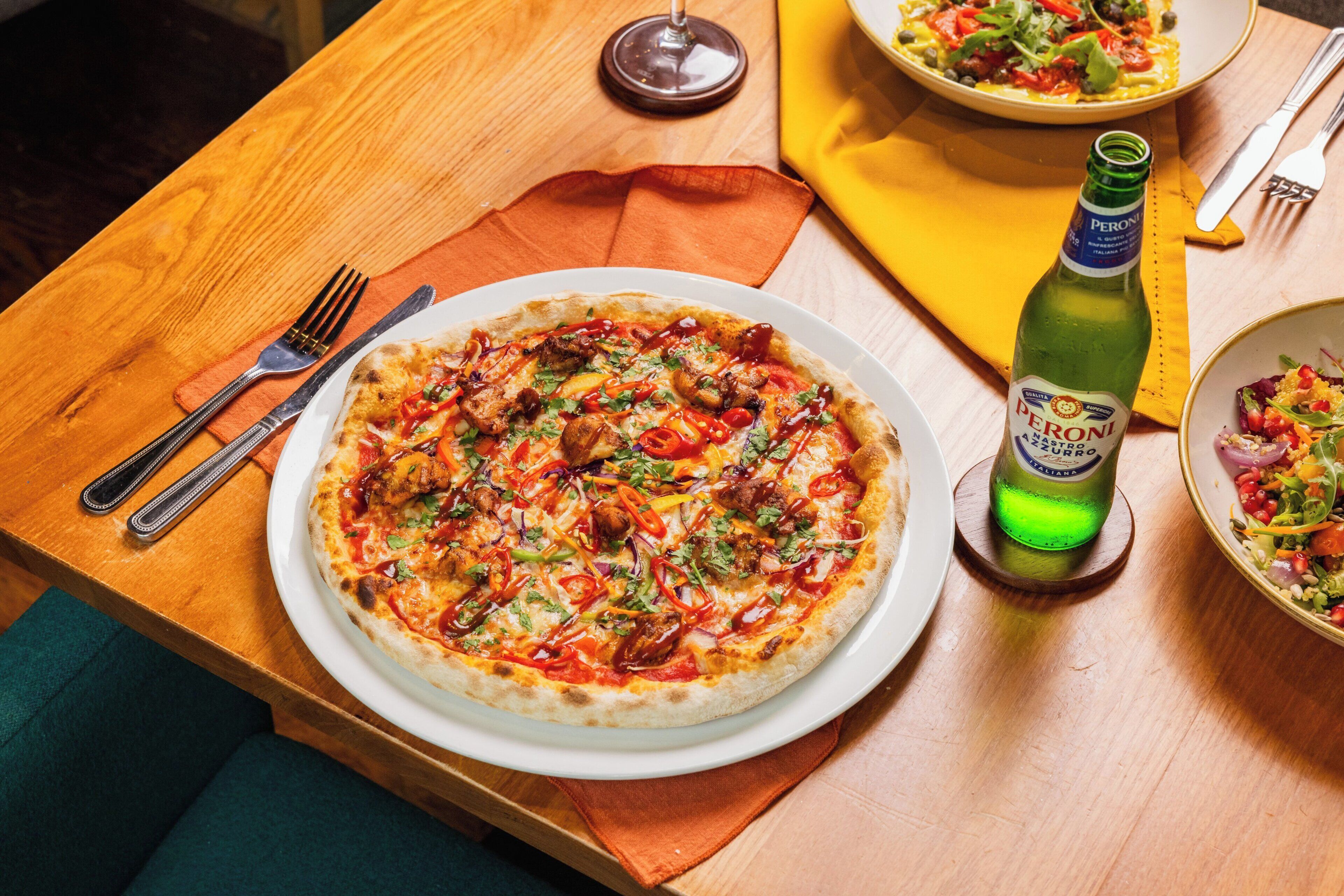 A pizza on a white plate with a bottle of Peroni Cuppone beer beside it.