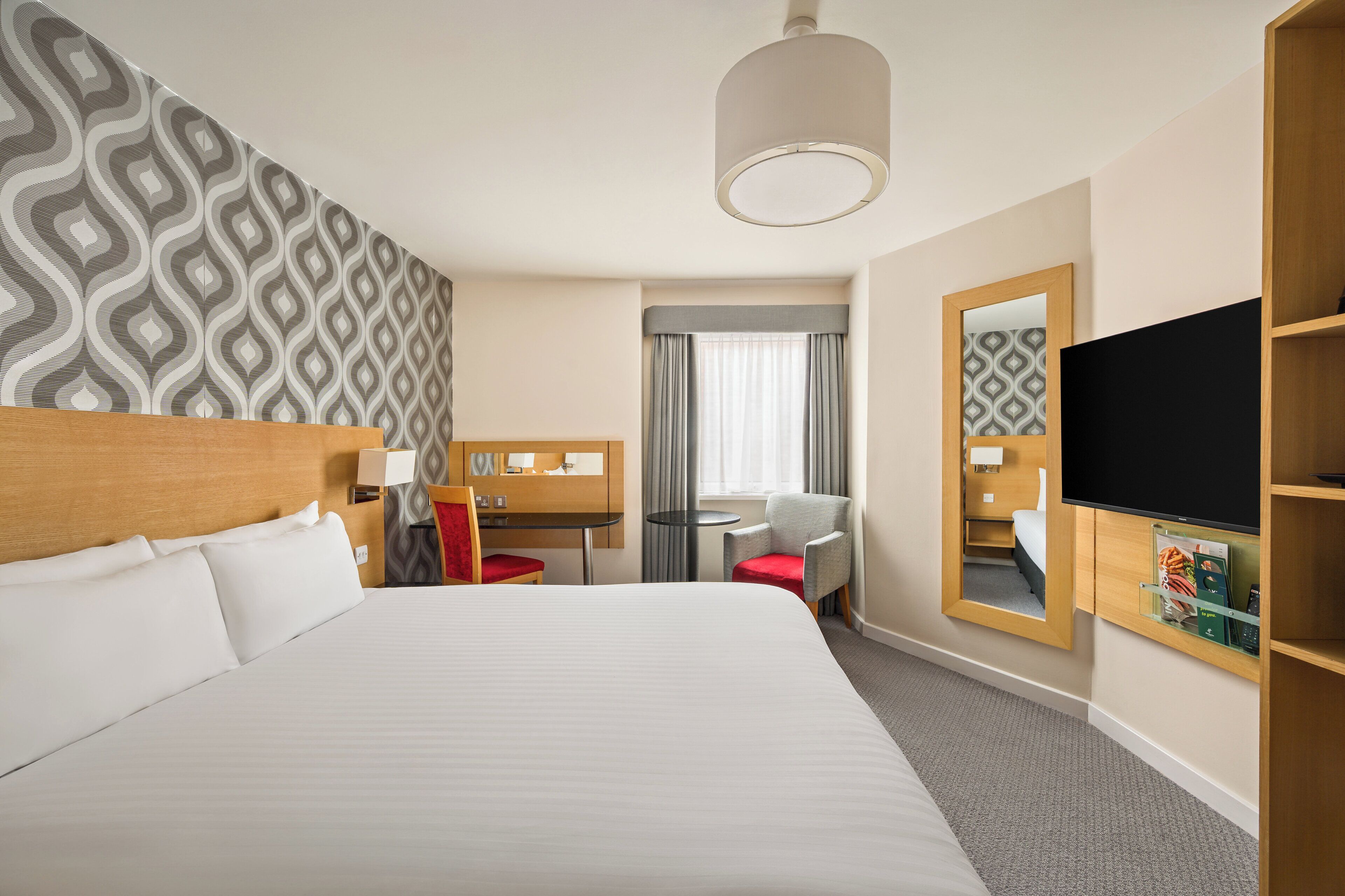 A hotel room with a large bed, a desk, a chair, a TV, and a mirror.