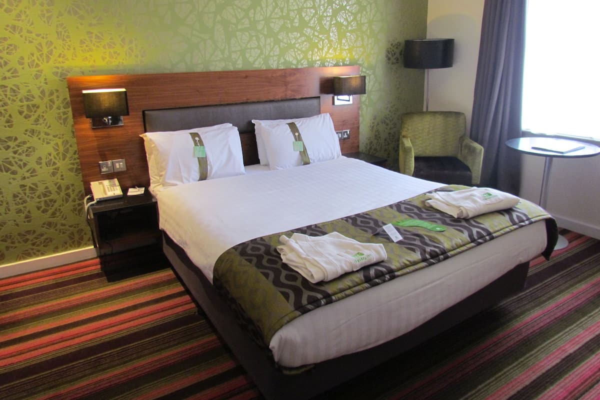 Habitación de hotel con una cama grande, una mesita de noche con teléfono, una silla, una pequeña mesa y una lámpara.