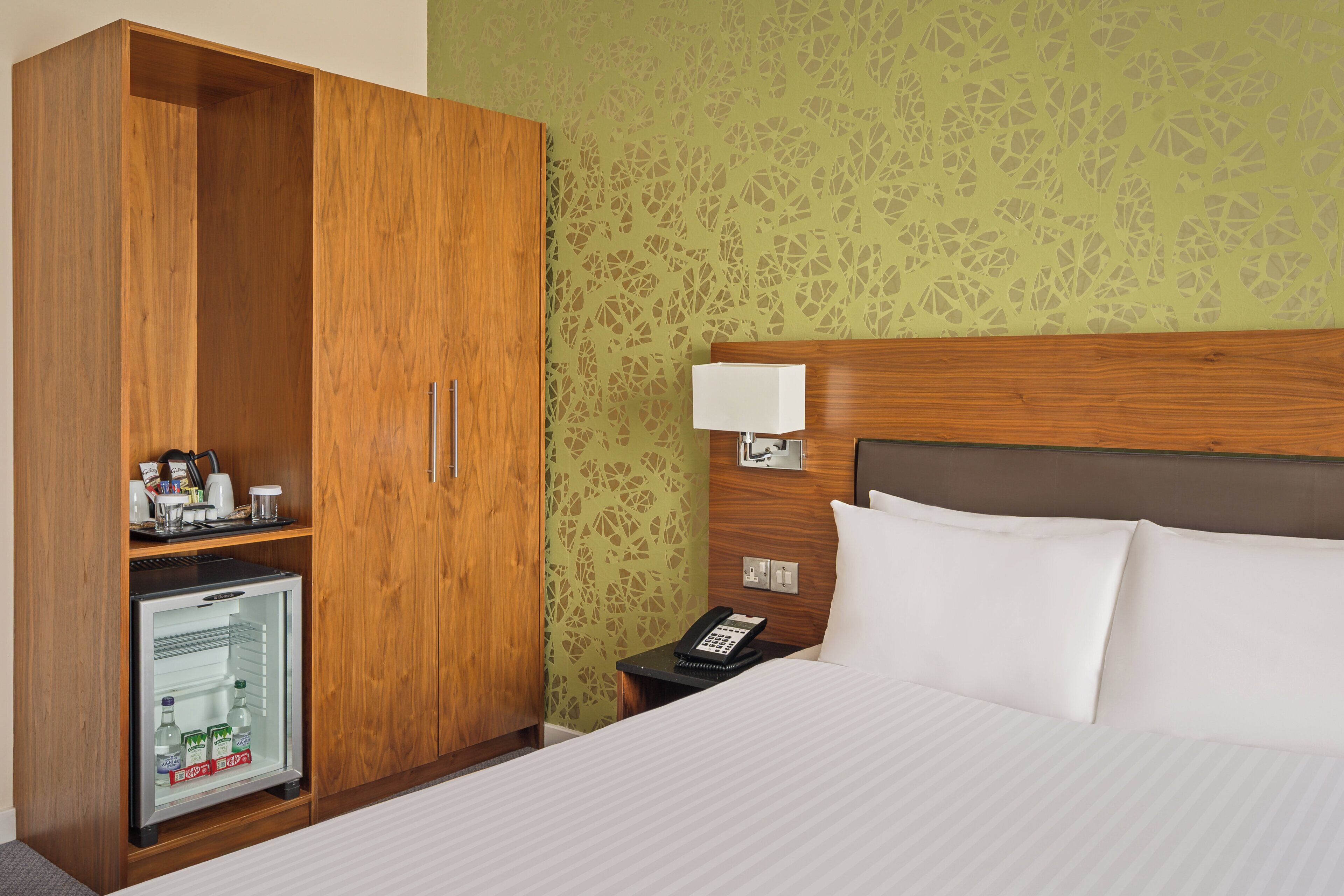 A hotel room with a bed, a wardrobe, a minibar, and a bedside table with a lamp and phone.