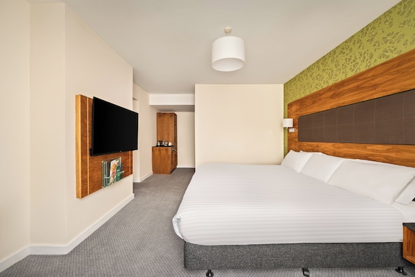 A hotel room with a large bed, a flat-screen TV mounted on the wall, a wooden headboard, and a small bedside table.