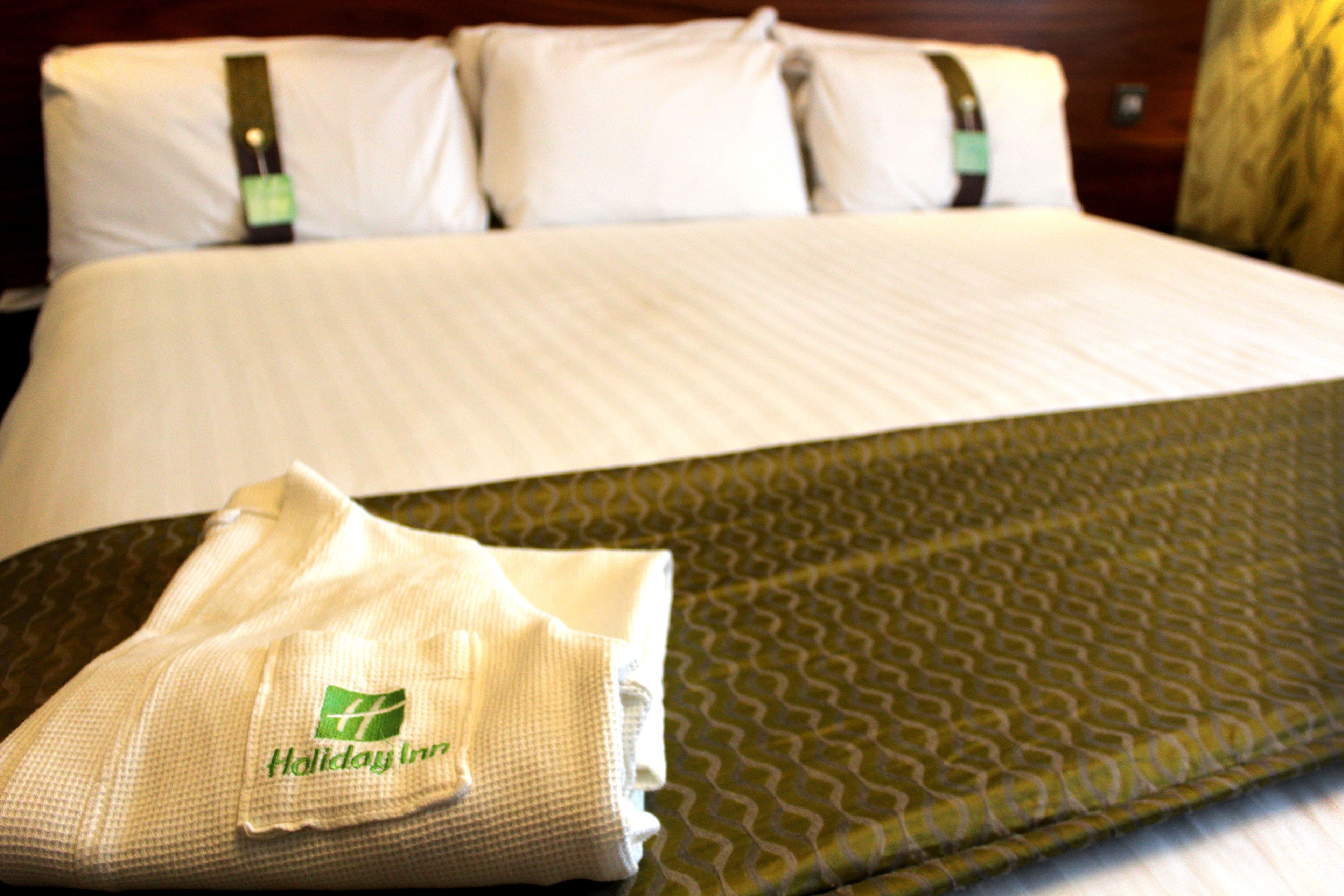 A neatly made bed with a green patterned bedspread and white pillows. A folded towel with a green logo is placed on the bed.