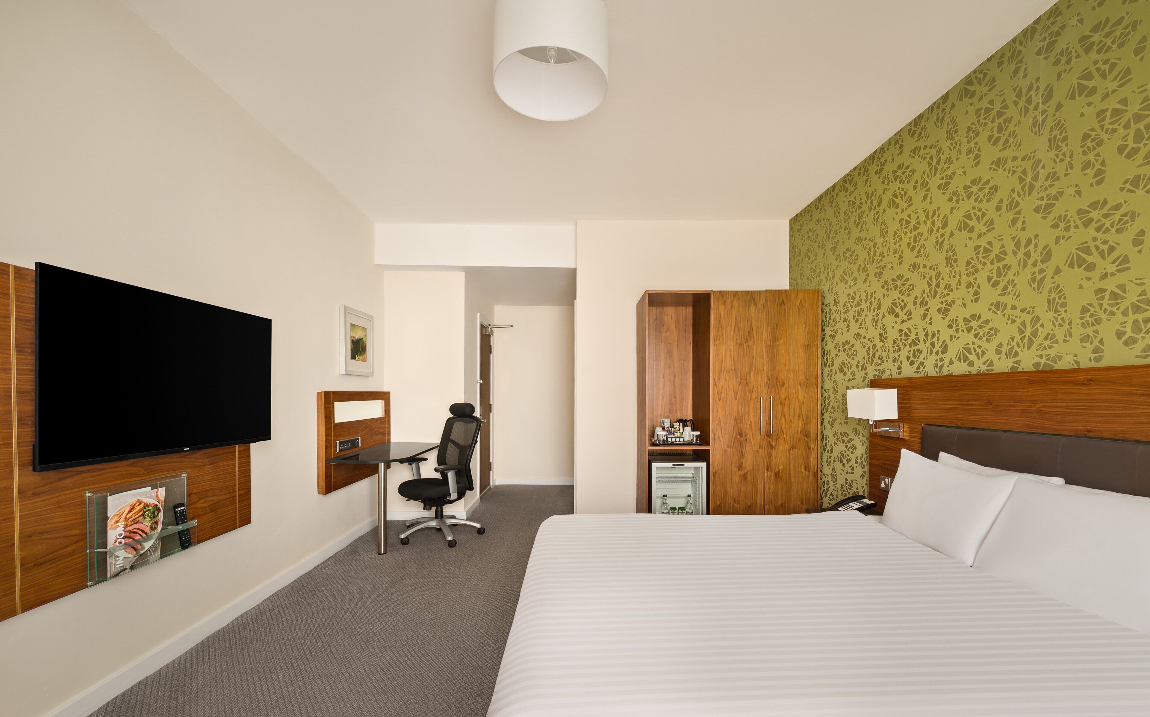 A hotel room with a large bed, a flat-screen TV, a desk with a chair, a wardrobe, and a small refrigerator.