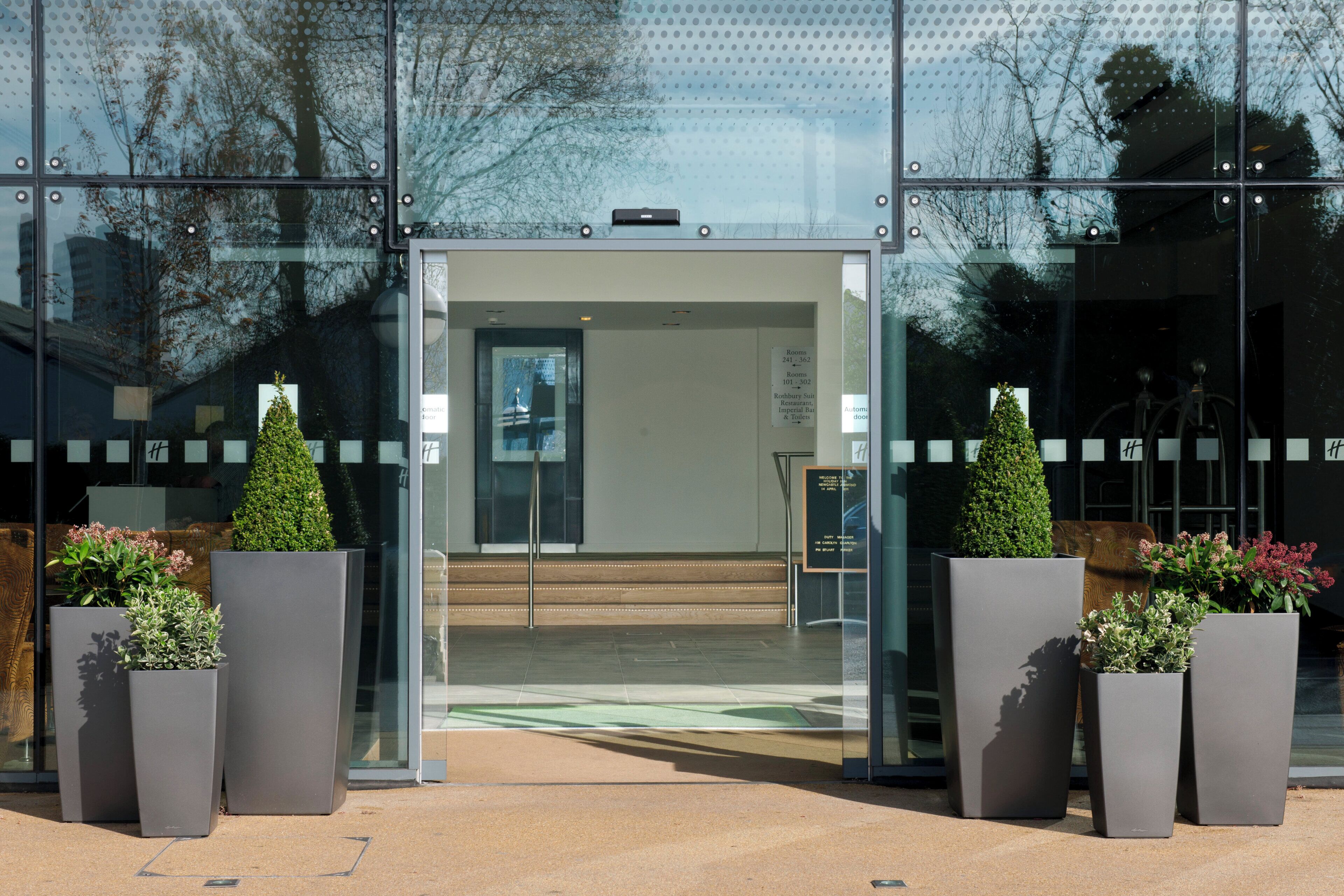 Une entrée moderne avec une porte en verre, des plantes en pot et un panneau indiquant les numéros des pièces.