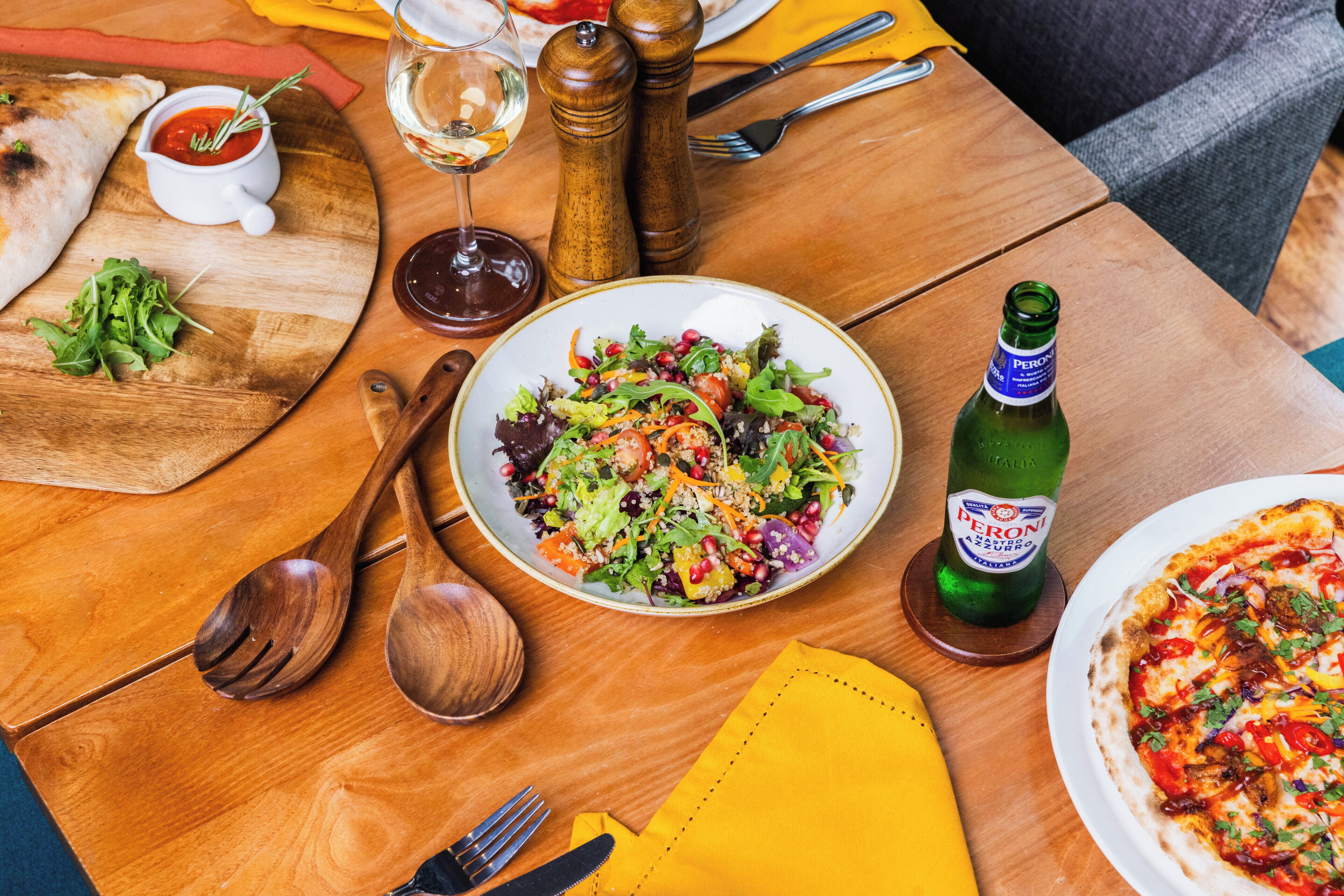 Een houten tafel met pizza, salade, wijn en een fles Peroni bier.