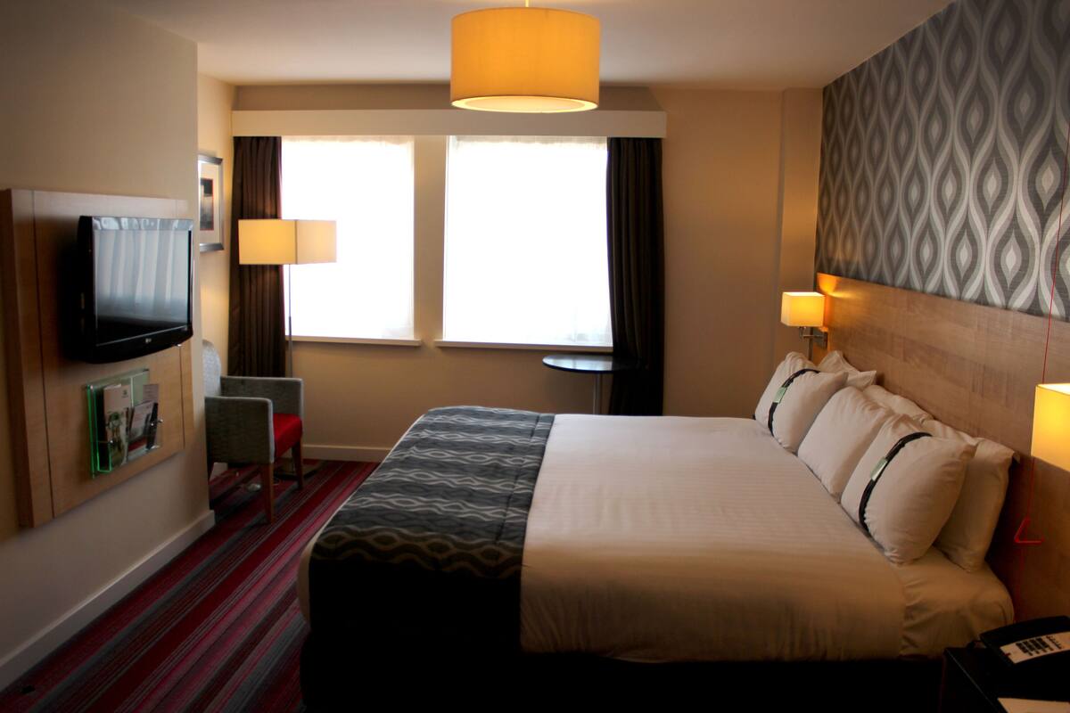 Habitación de hotel con una cama grande, dos lámparas de noche, un televisor de pantalla plana, una mesita y un ventanal con cortinas.